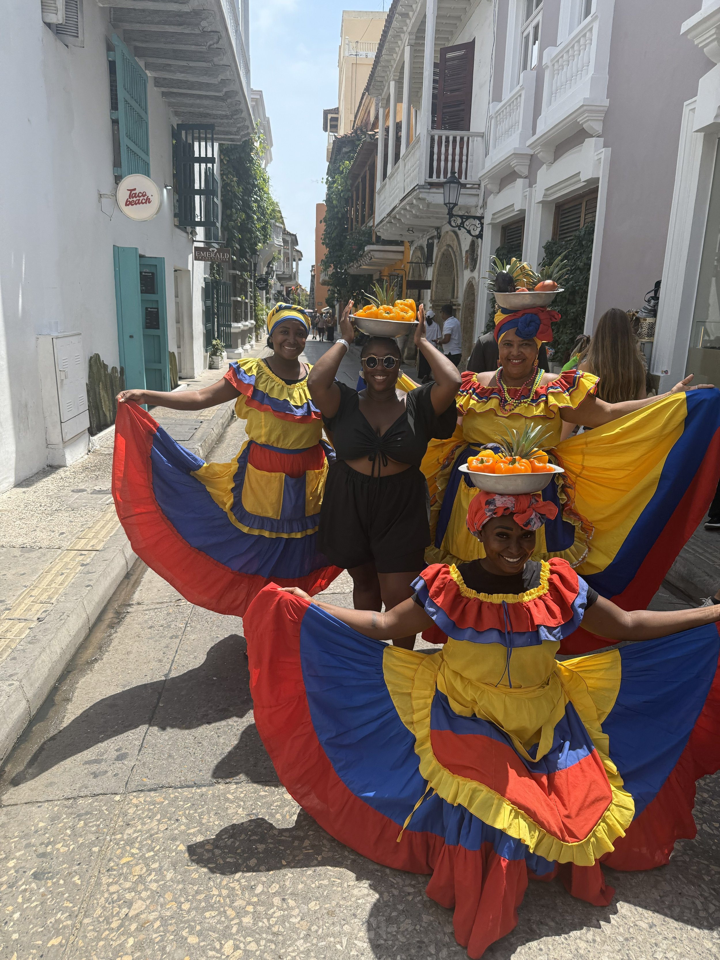 Four women dressed in colorful traditional Colombian outfits, holding bowls of fruit on their heads, dancing in a street with colonial-style buildings.