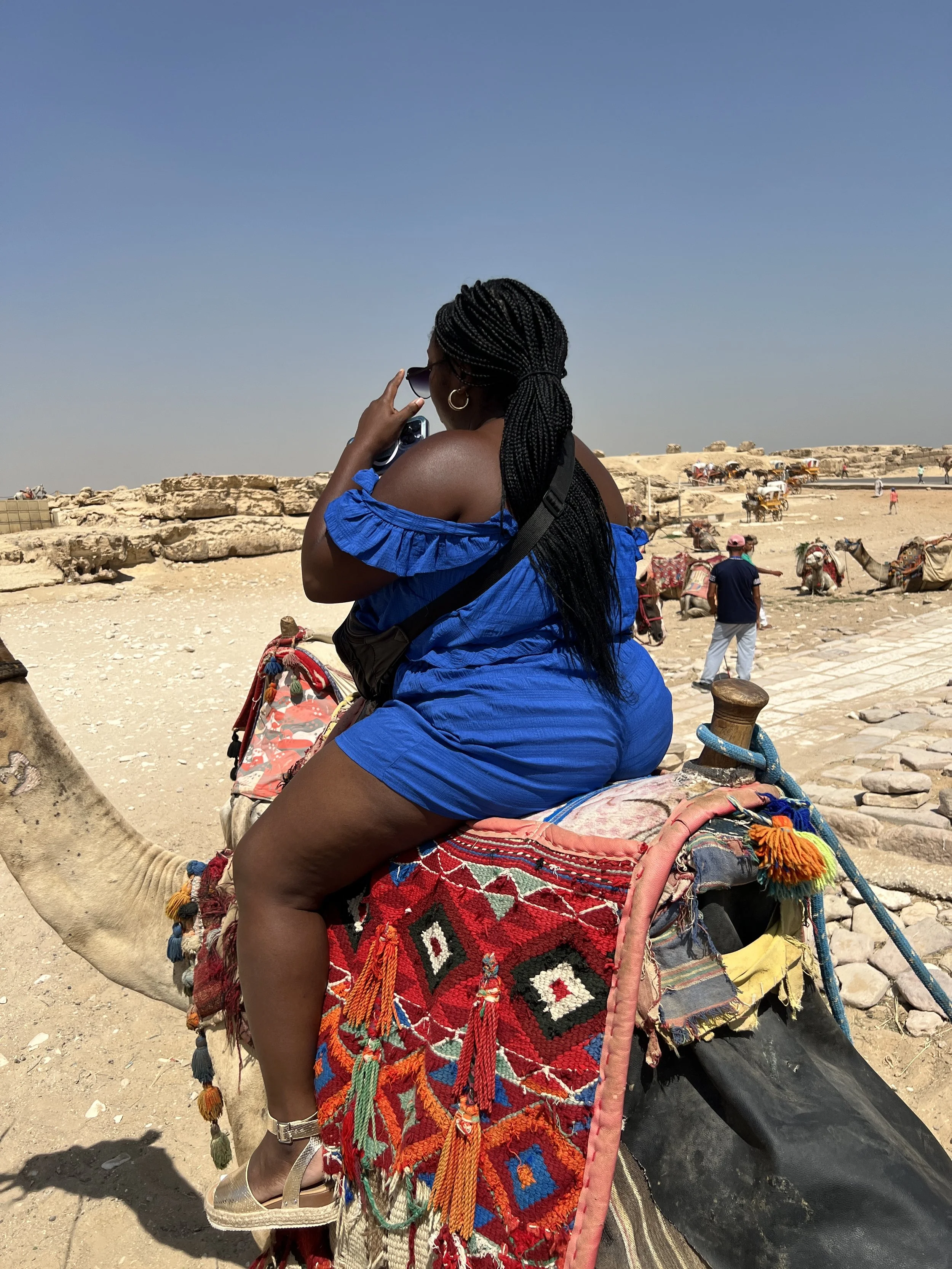 Latrice in a blue outfit sitting on a camel with a colorful saddle, talking on her phone, with a desert landscape and other camels in the background.