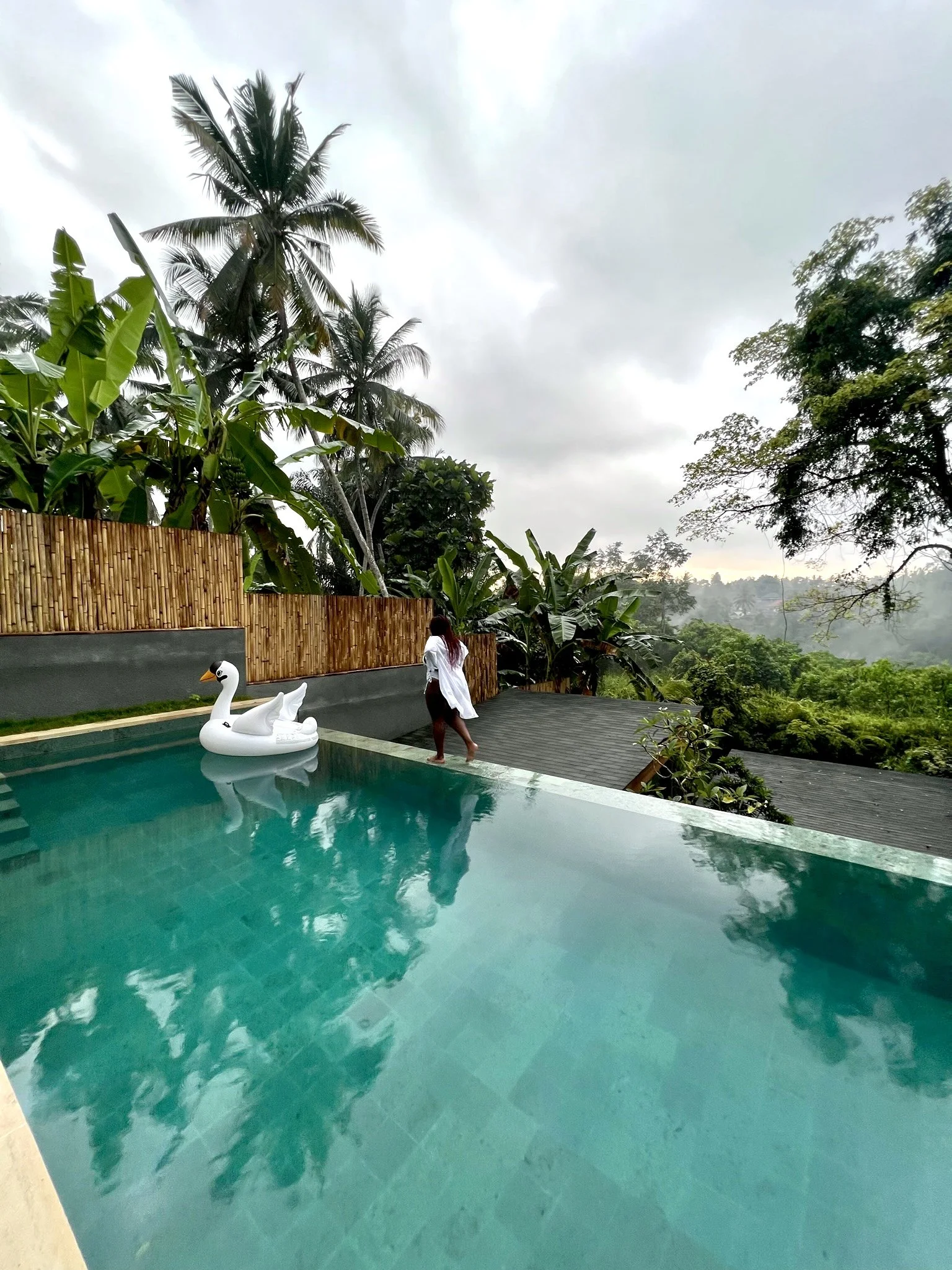 Latrice stands at the edge of an infinity pool overlooking a lush, tropical landscape with palm and banana trees, under a cloudy sky.