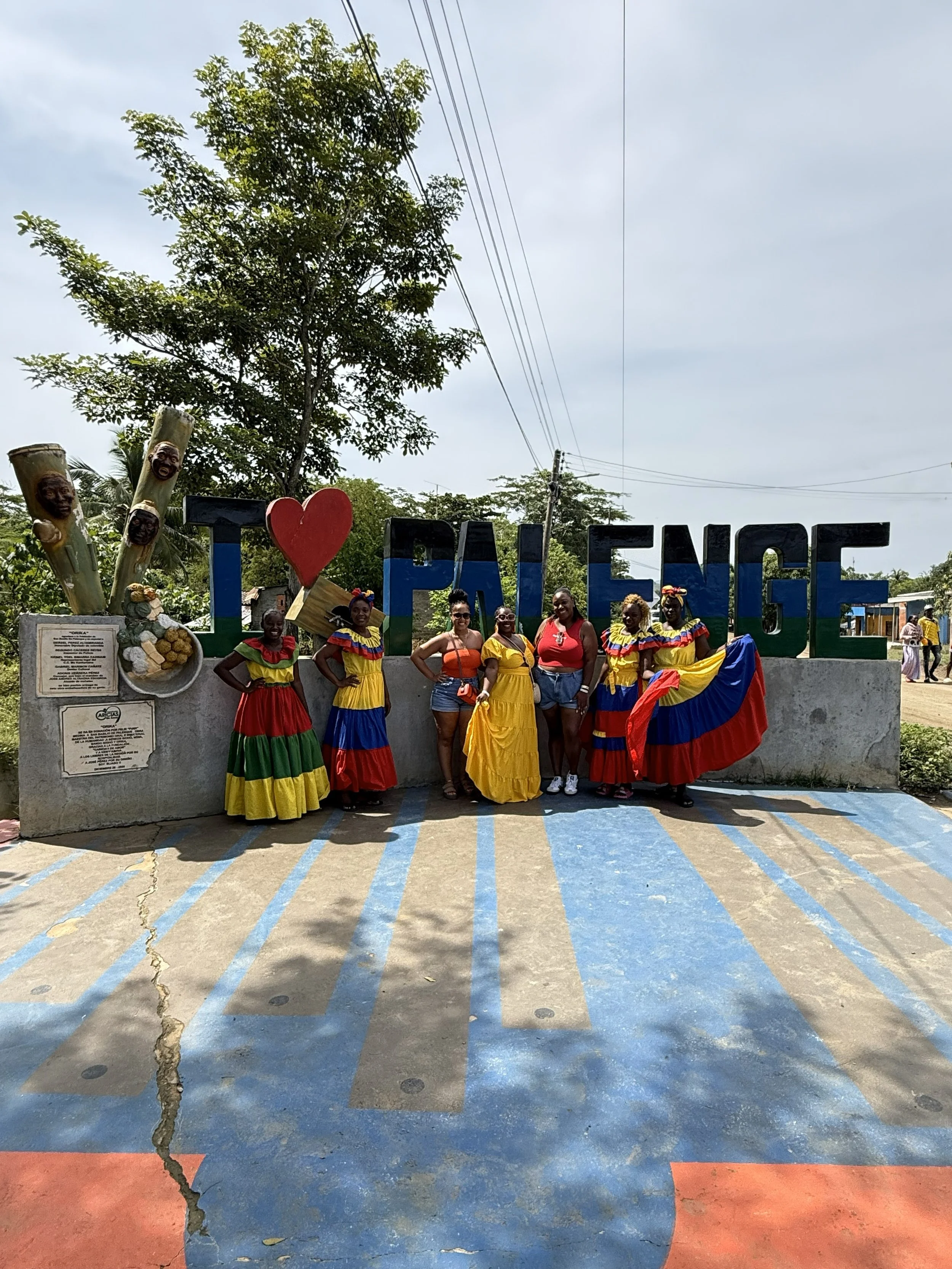 Group of women in colorful traditional dresses standing in front of a large sign that reads 'I love PALIENCE' in an outdoor setting with trees and power lines overhead.