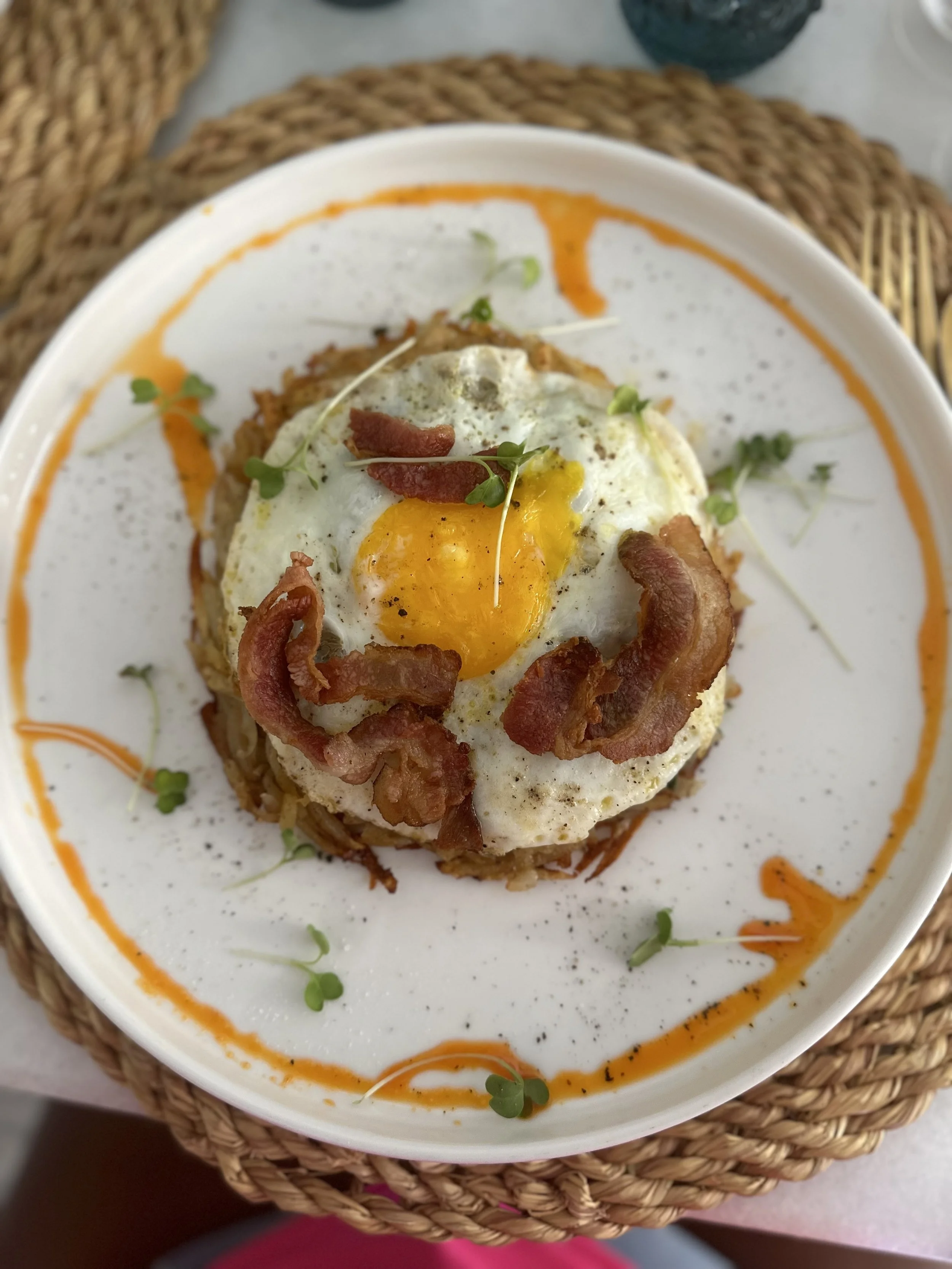 A plate of huevos motuleños, topped with a fried egg, bacon, and microgreens, served with a creamy sauce with a swirl of orange sauce on the rim of the dish.