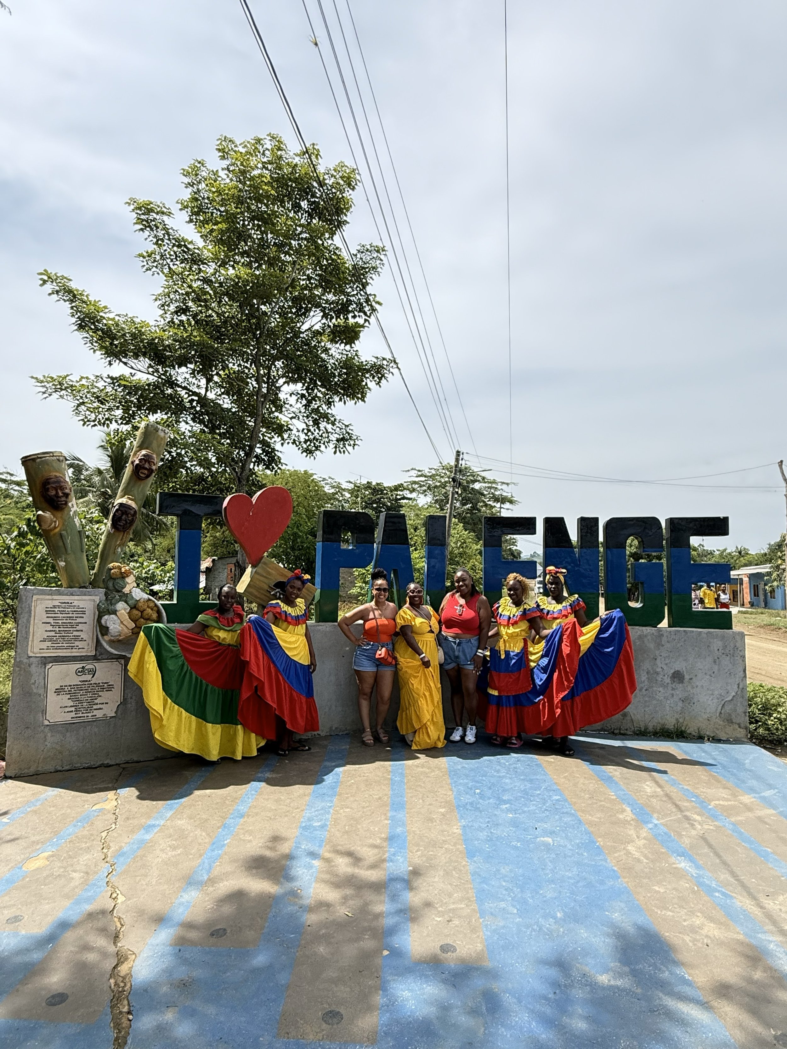 Group of six women standing in front of a large sign that reads "I LOVE PALENQUE," with colorful dresses and traditional attire, under a cloudy sky with trees and power lines in the background.