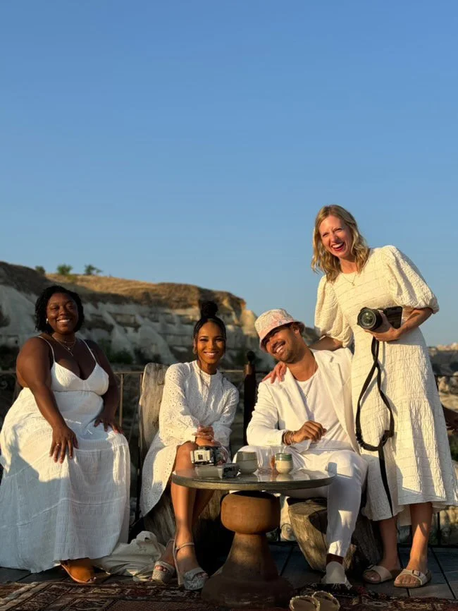 Four diverse friends enjoying a sunny outdoor gathering, with a rocky landscape and clear blue sky in the background. One woman standing on the right holds a camera, and another woman sitting in the middle has a vintage camera on her lap. They are al