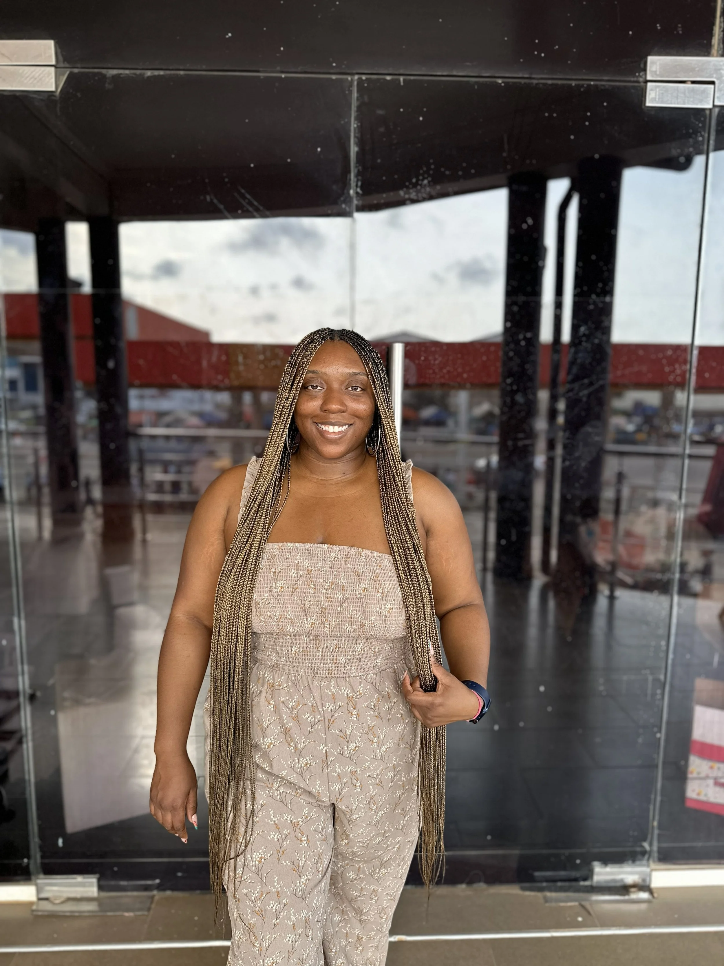 Latrice with long braided hair and hoop earrings, wearing a beige patterned strapless top and matching pants, standing outside a glass building with a parking lot and cloudy sky in the background, smiling at the camera.