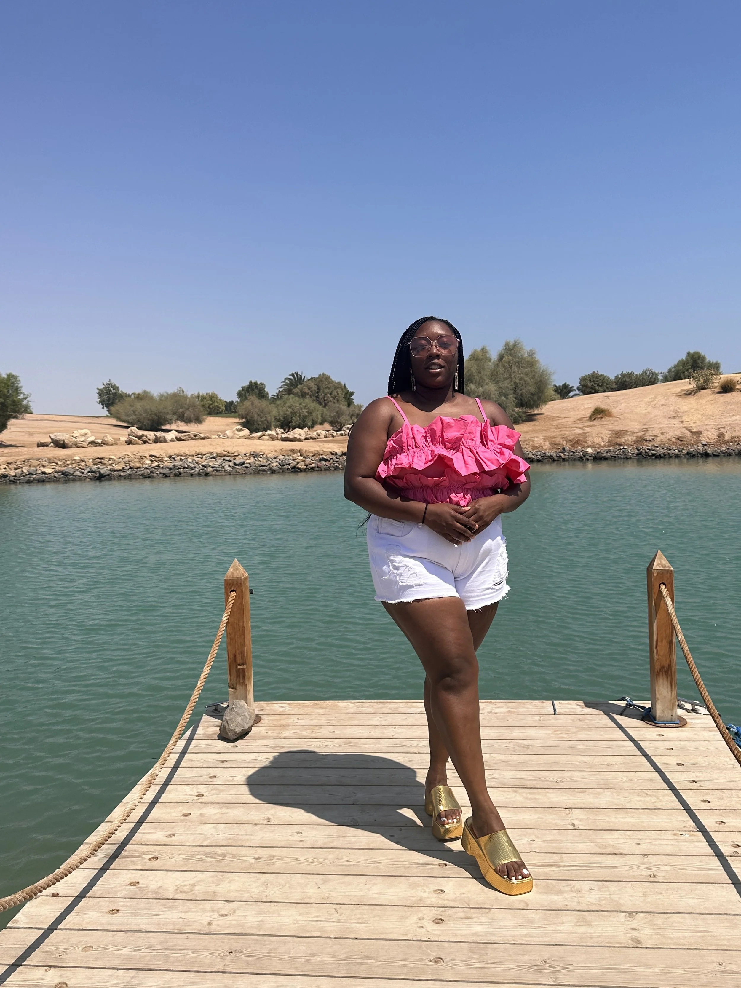 Latrice standing on a wooden dock by a body of water, wearing a pink ruffled top, white shorts, and yellow platform sandals, with trees and a sandy landscape in the background under a clear blue sky.