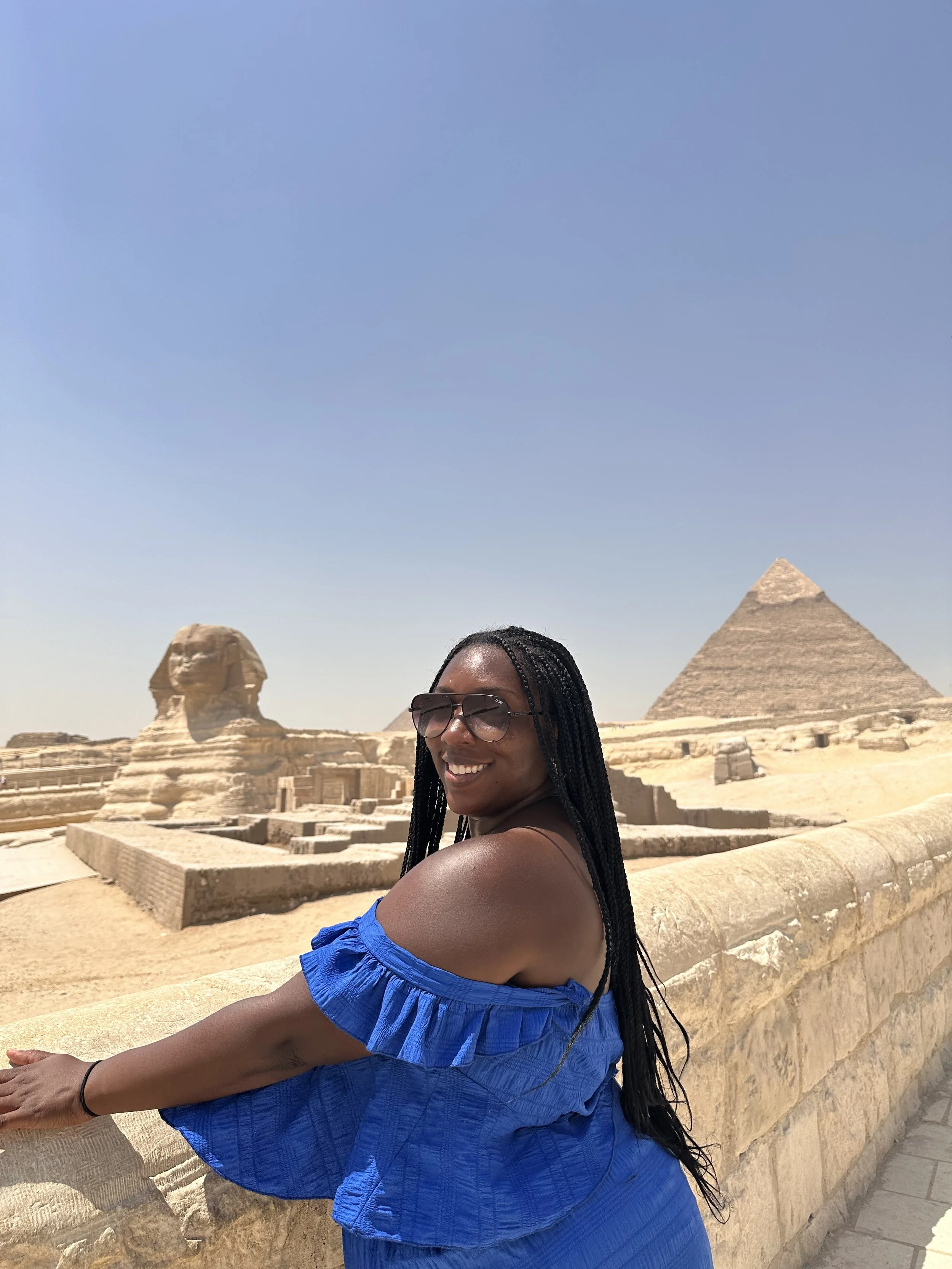 Latrice wearing sunglasses and a blue off-shoulder dress standing on a stone platform with the Great Pyramid of Giza and the Sphinx in the background during daytime.