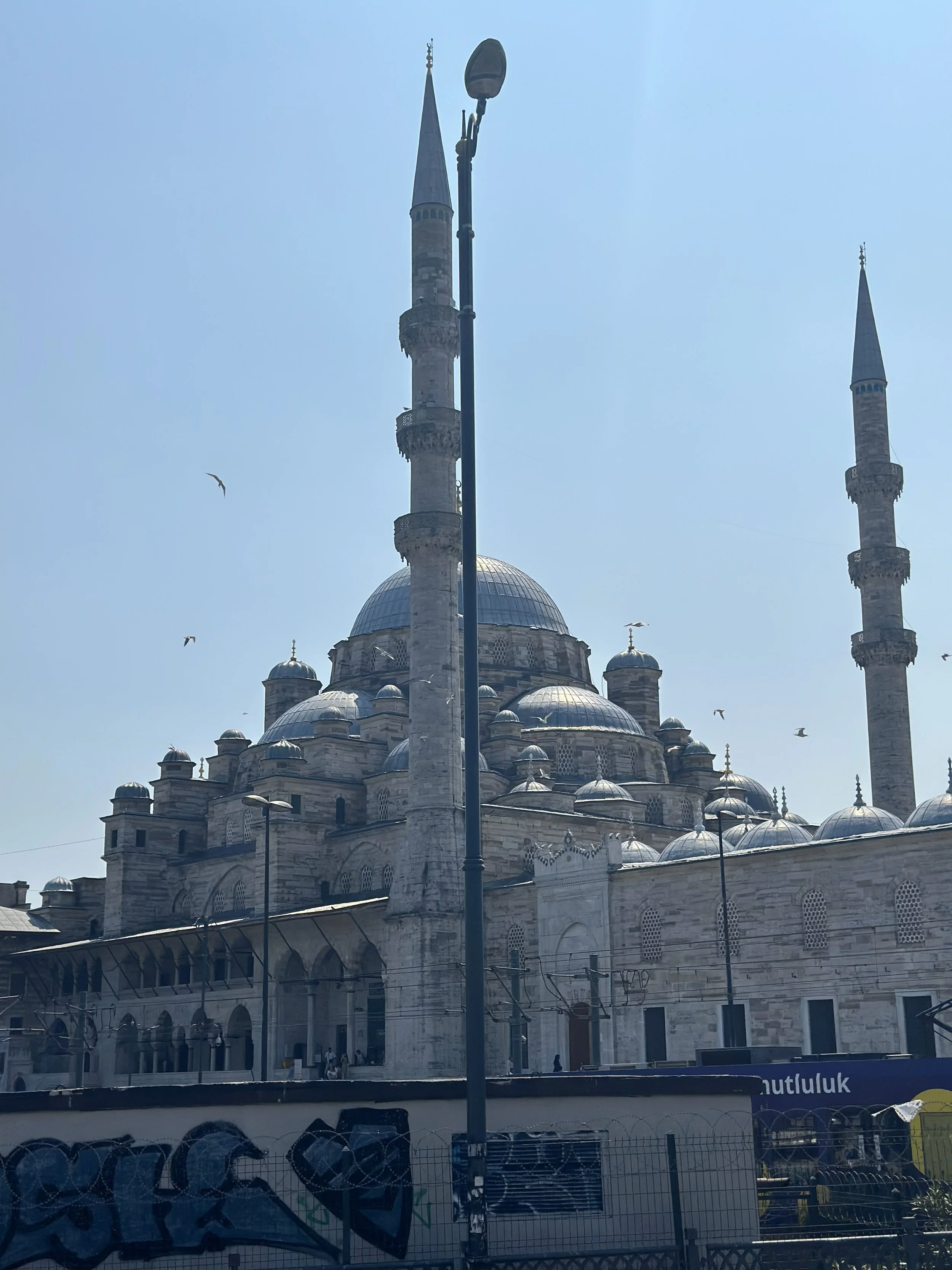 A mosque with multiple domes and four minarets, taken on a clear, sunny day with a light blue sky and some birds flying nearby.