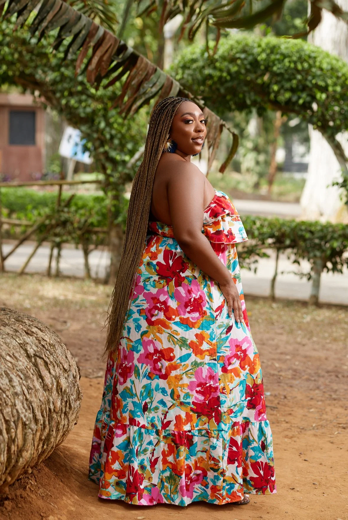 Latrice with long braided hair wearing a colorful floral strapless dress standing outdoors in front of green bushes and trees.