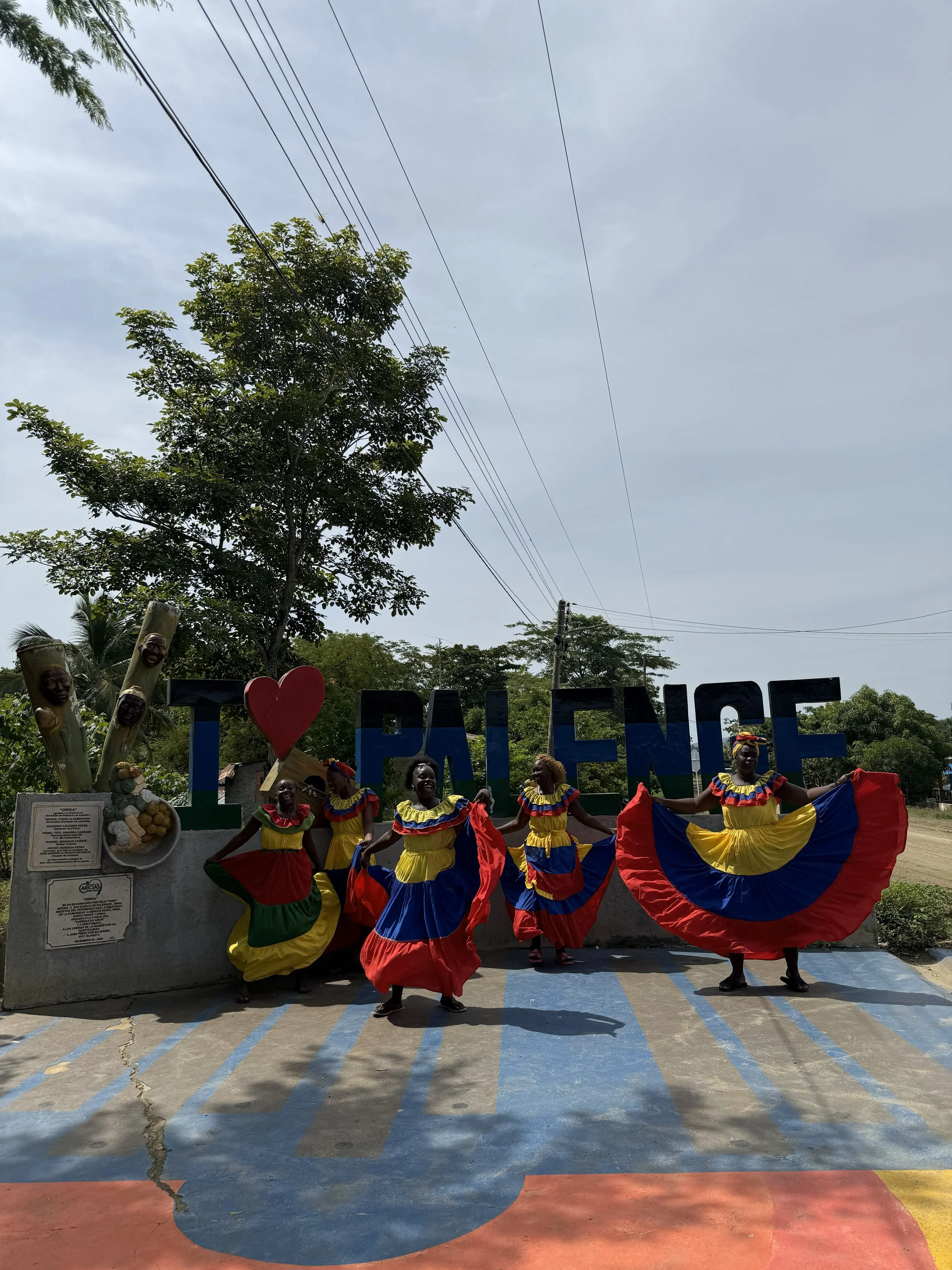 Group of women in colorful traditional dresses performing a dance in front of a sign that says 'I ❤️ Palenque', surrounded by trees and outdoor setting.