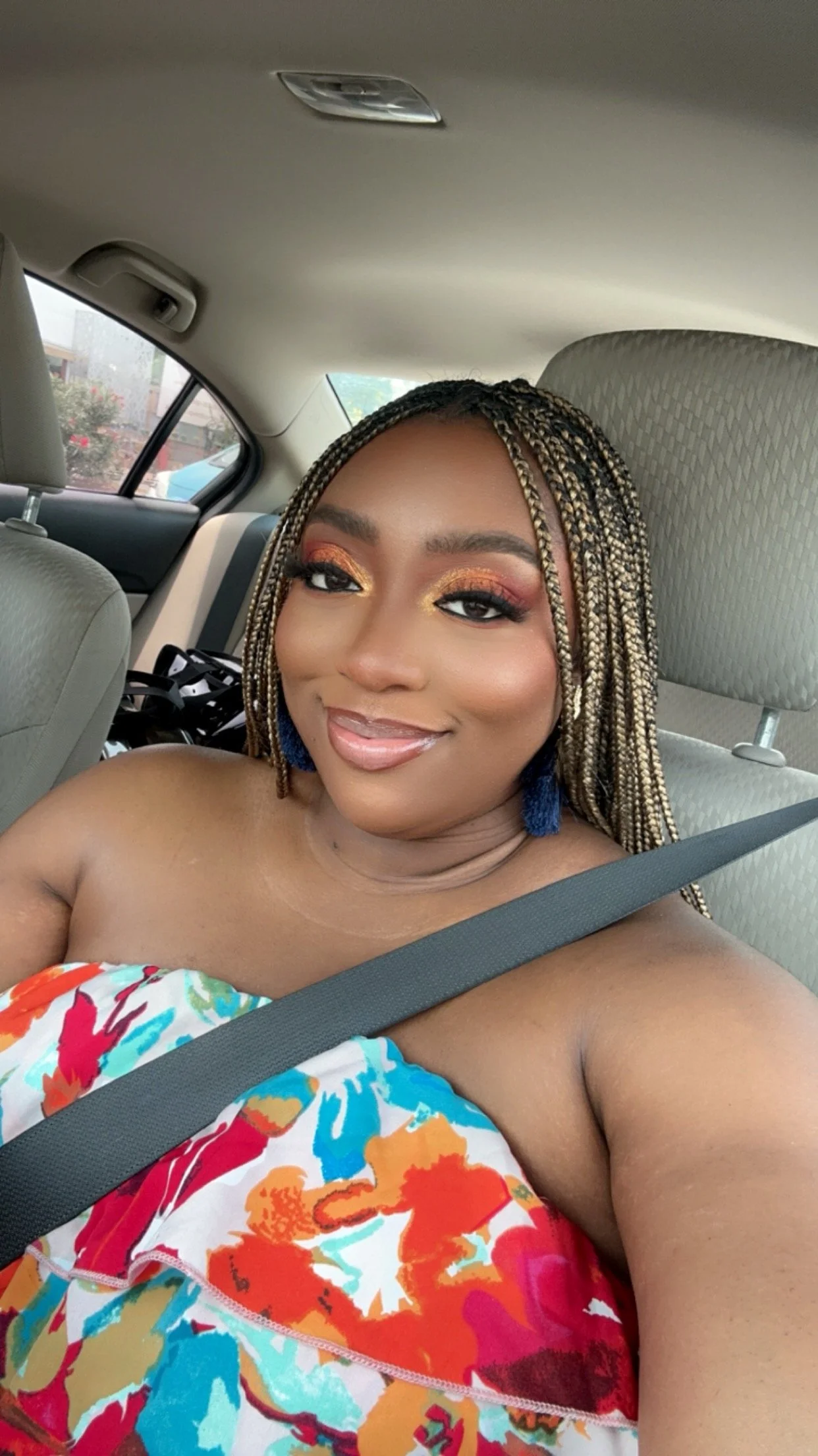 Latrice with braided hair, colorful makeup, and blue earrings, sitting in a car wearing a strapless floral dress and smiling at the camera.