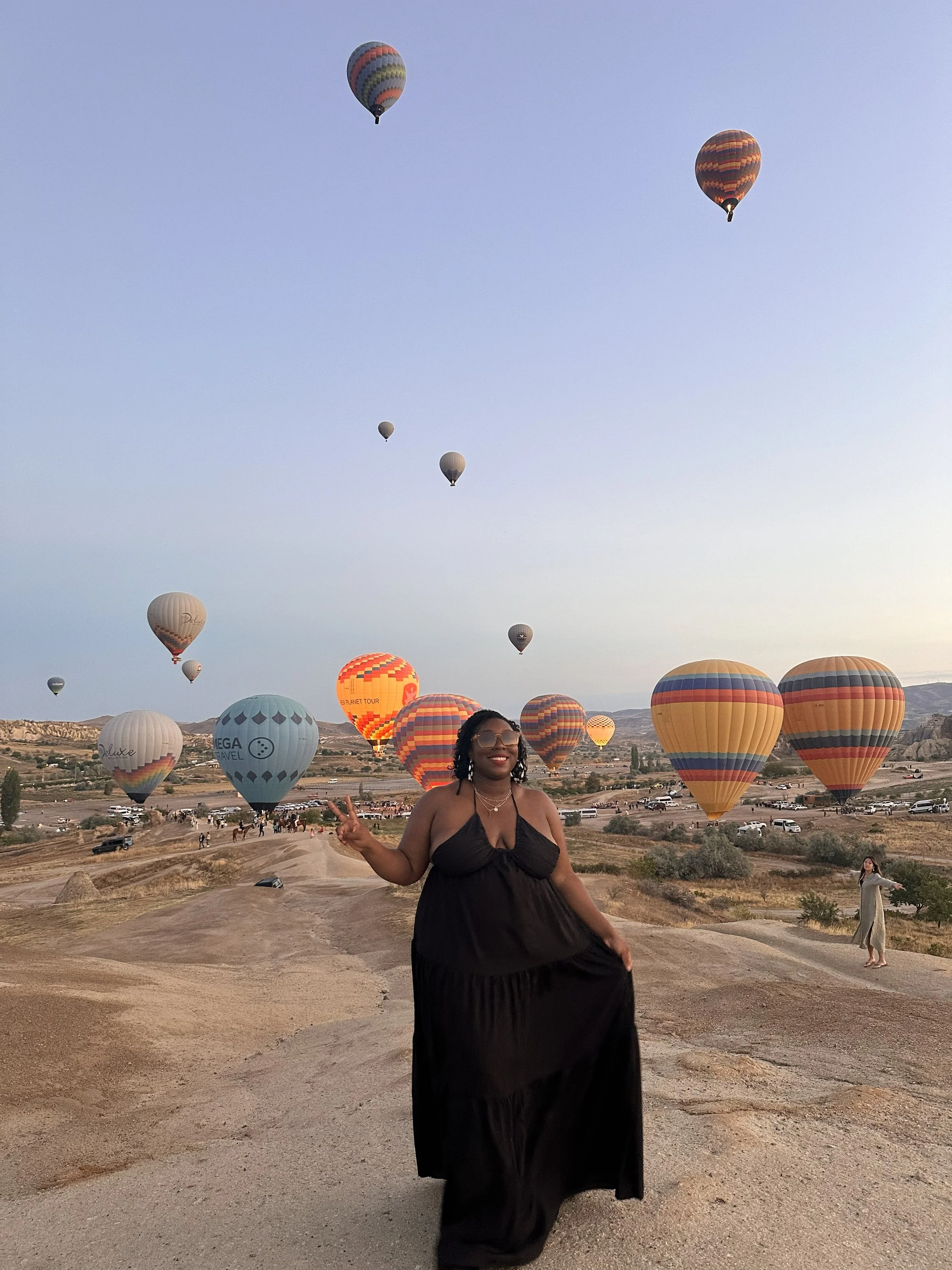 Latrice in a black dress smiling and making a peace sign at a hot air balloon festival, with numerous colorful hot air balloons in the sky and on the ground.