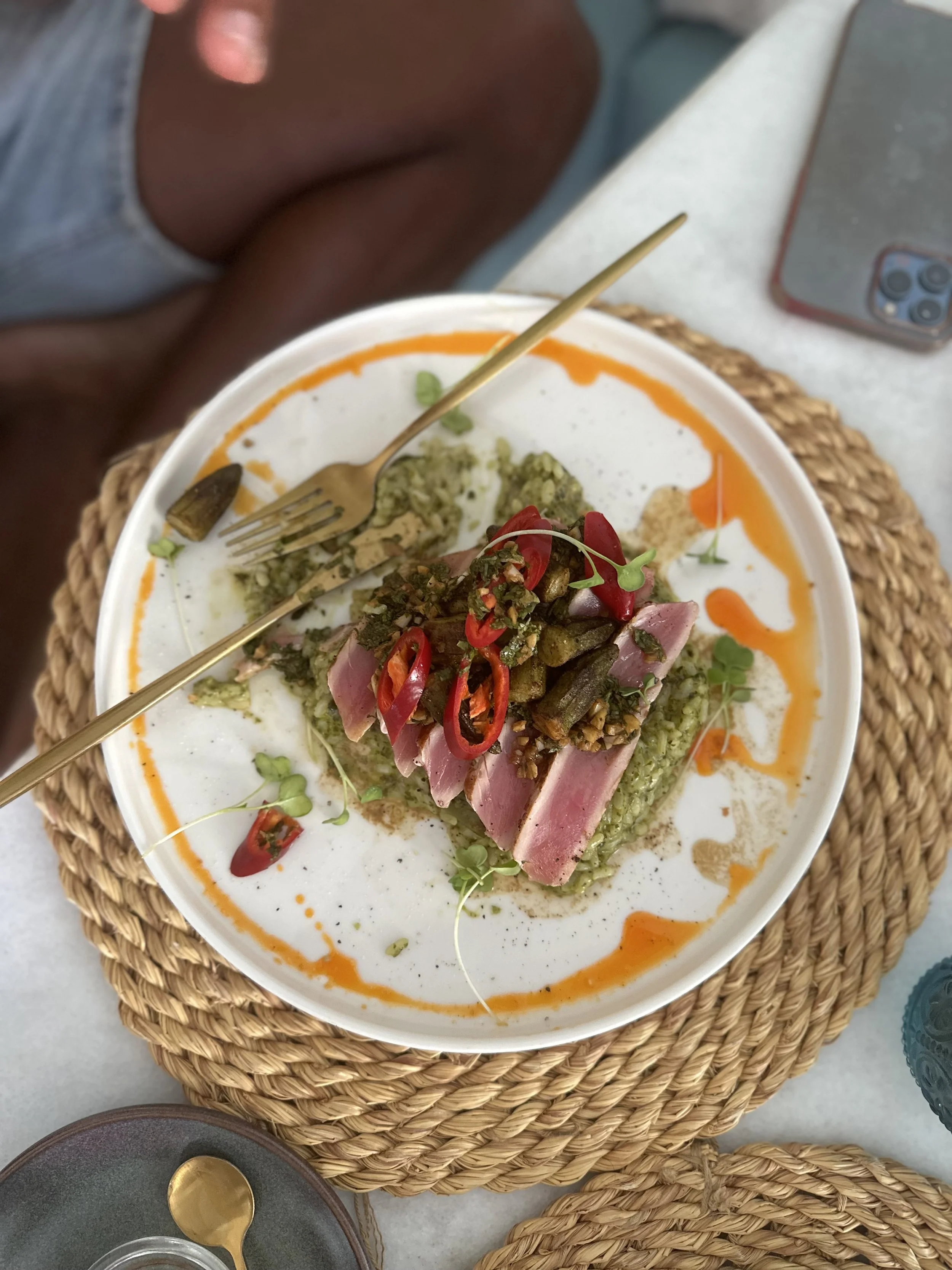 A bowl of food with sliced pink meat, sauces, chopped vegetables, and red sliced chili peppers on a white plate on a woven placemat.