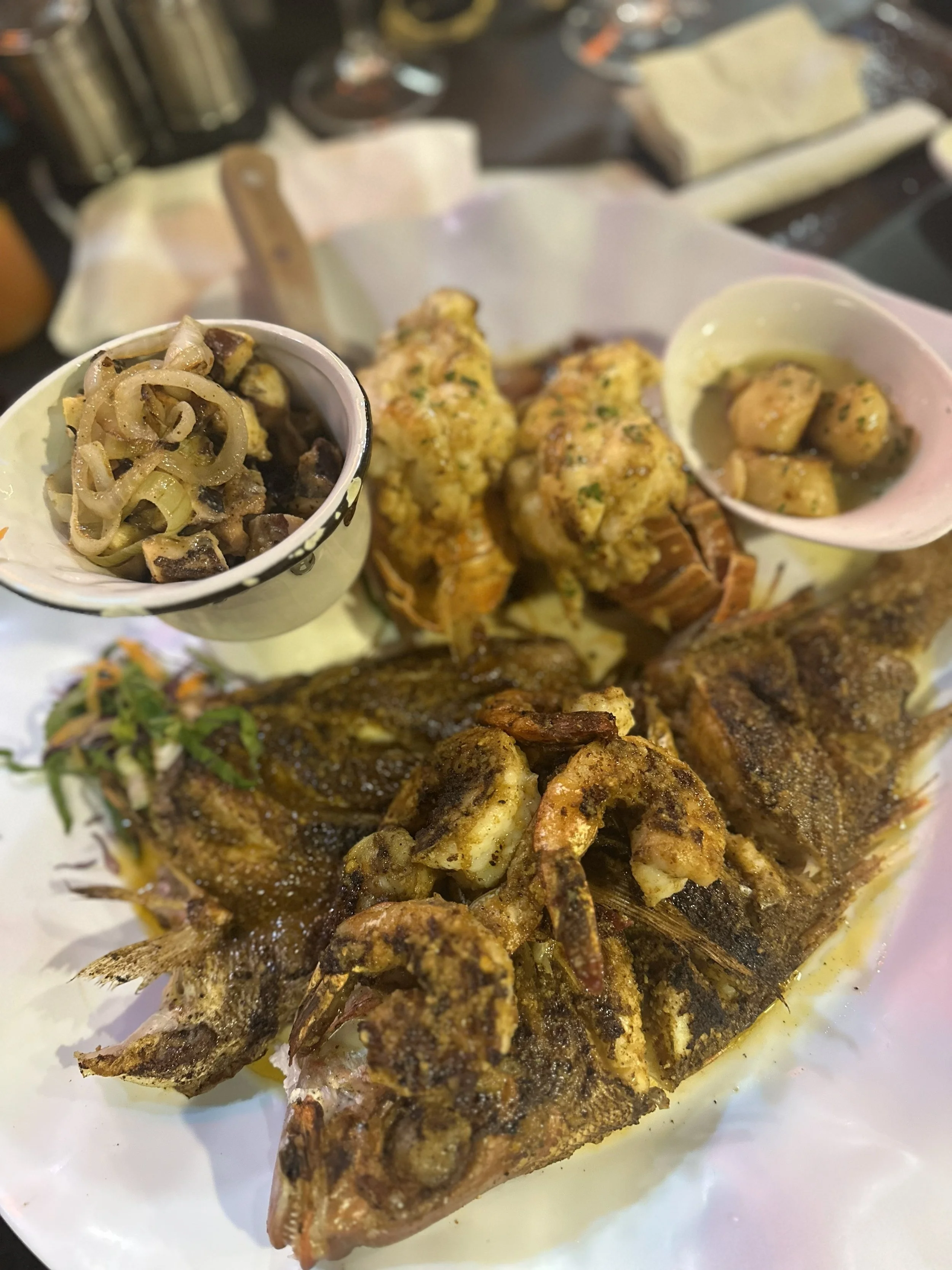 Fried fish with shrimp, boiled potatoes, grilled chicken, sautéed vegetables, and small bowls of sides on a white plate.
