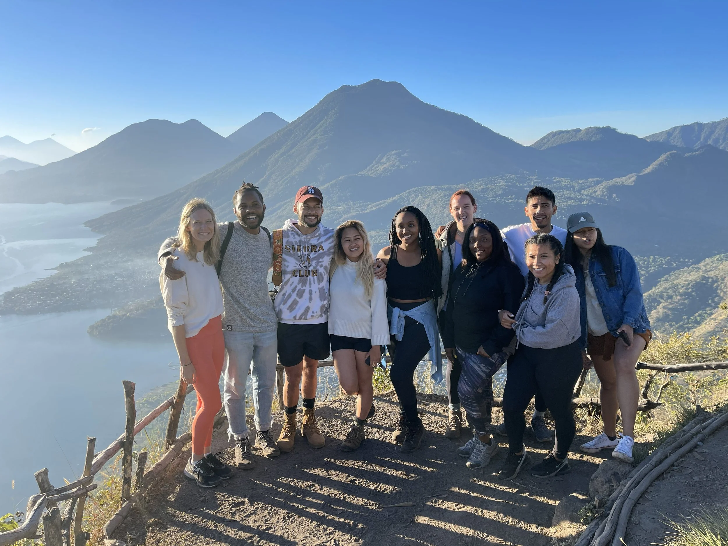 Group of eleven diverse young adults standing together on a mountain trail with breathtaking views of mountains and a lake in the background, celebrating outdoors.
