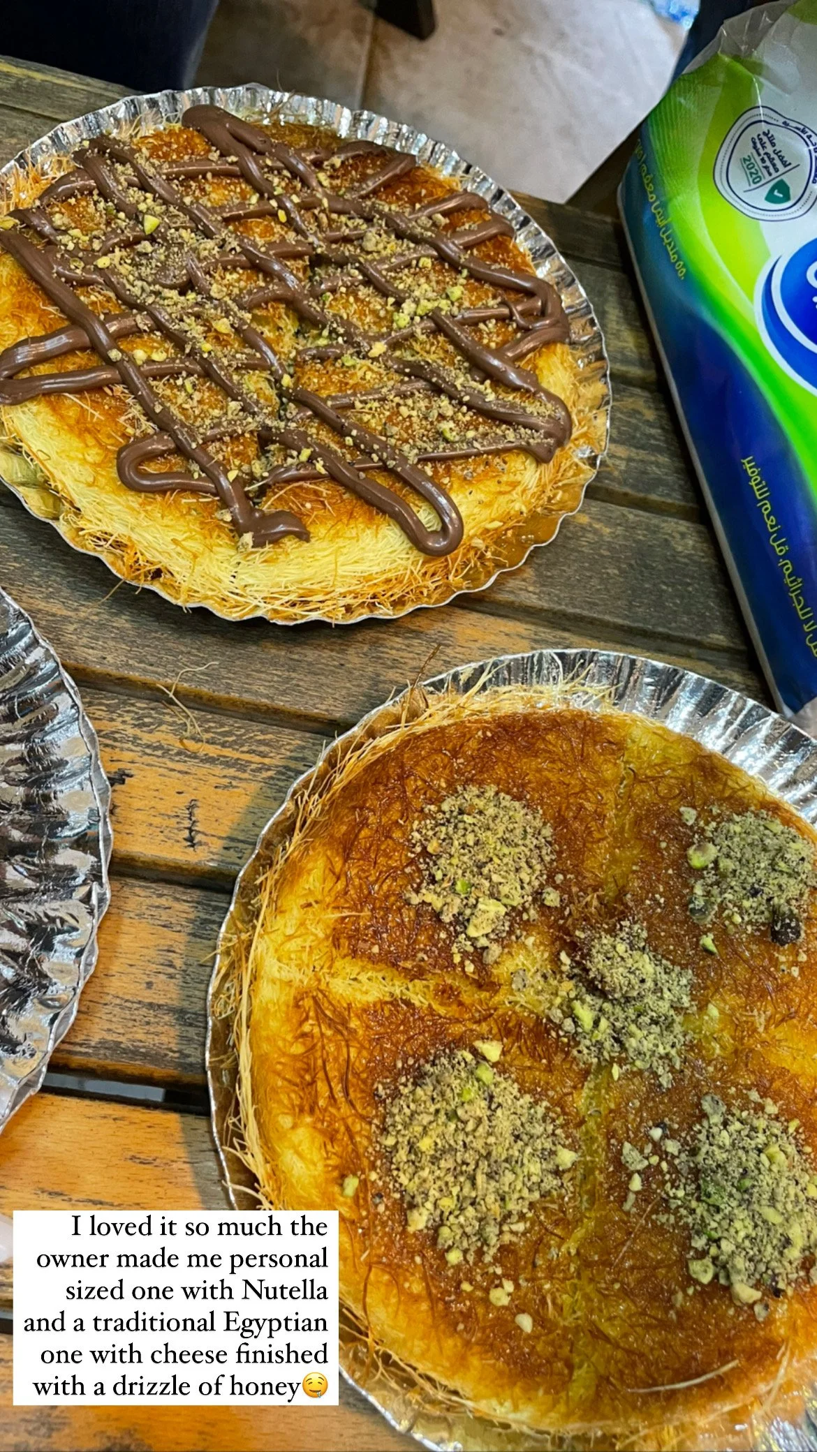 Two trays of Egyptian desserts, one with Nutella topped with crushed pistachios and honey, and the other with shredded phyllo dough drizzled with honey and crushed pistachios, on a wooden table.