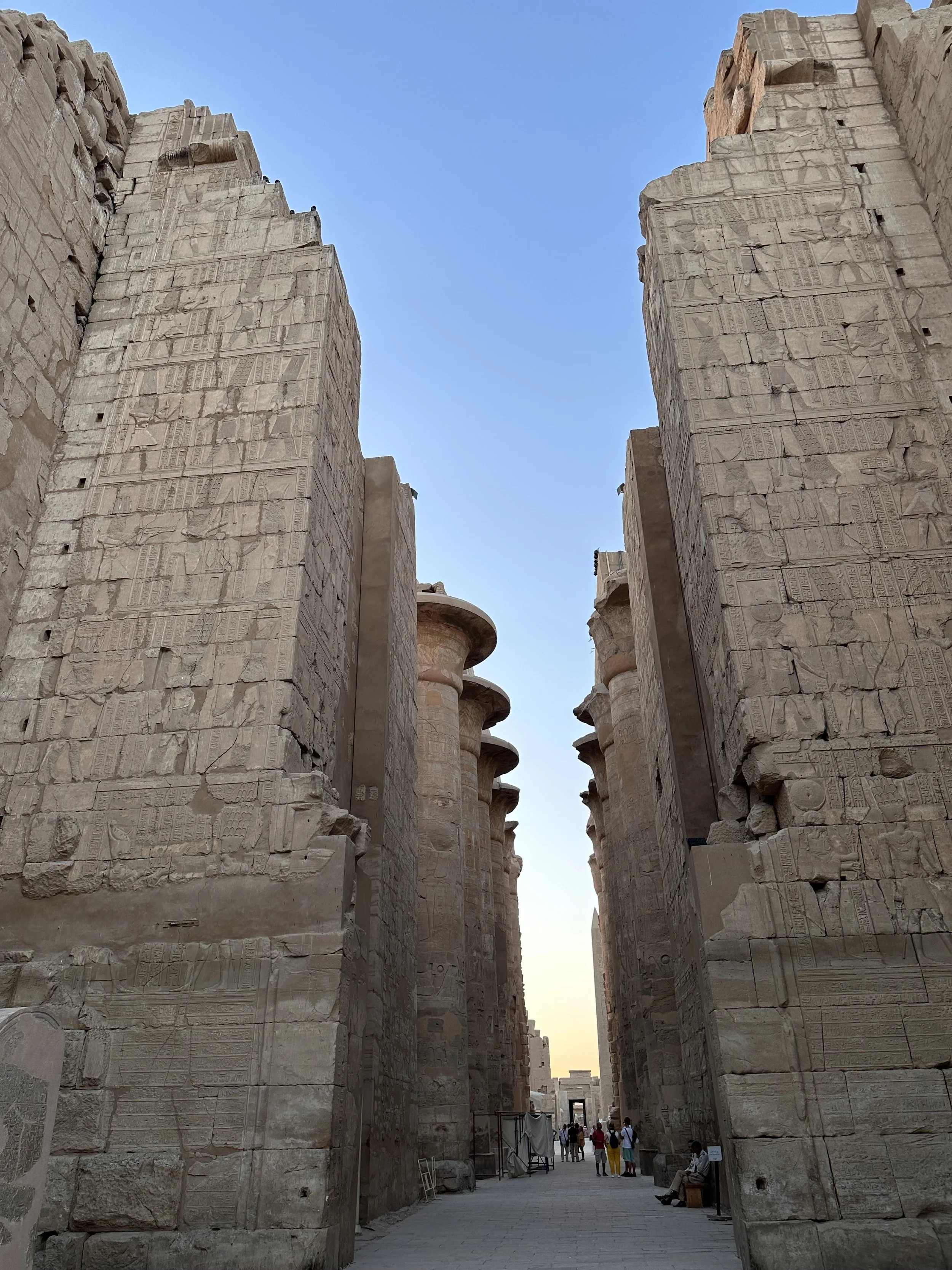 Ancient Egyptian temple ruins with large stone walls and columns, with visitors walking through the temple entrance during the daytime.