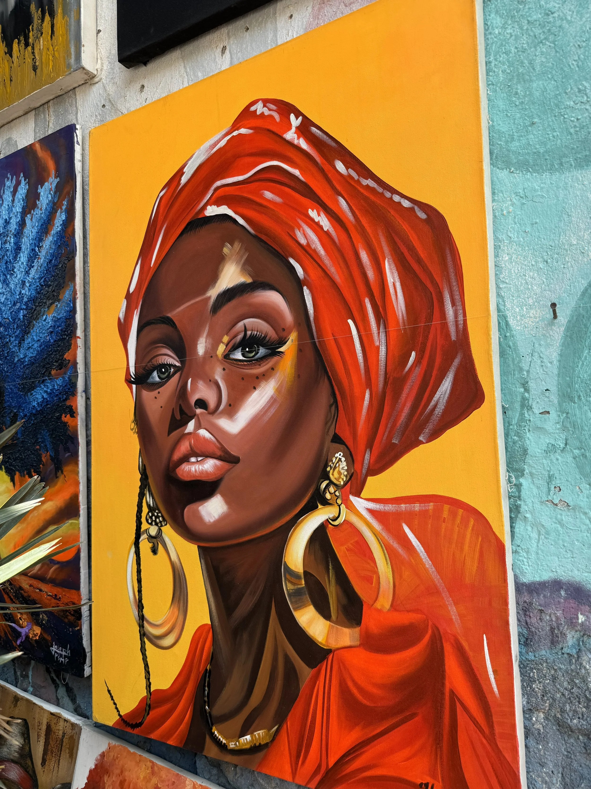 A portrait mural of a woman with dark skin, wearing large gold hoop earrings, a red headwrap, and makeup on her lips and eyes, painted on a yellow background.