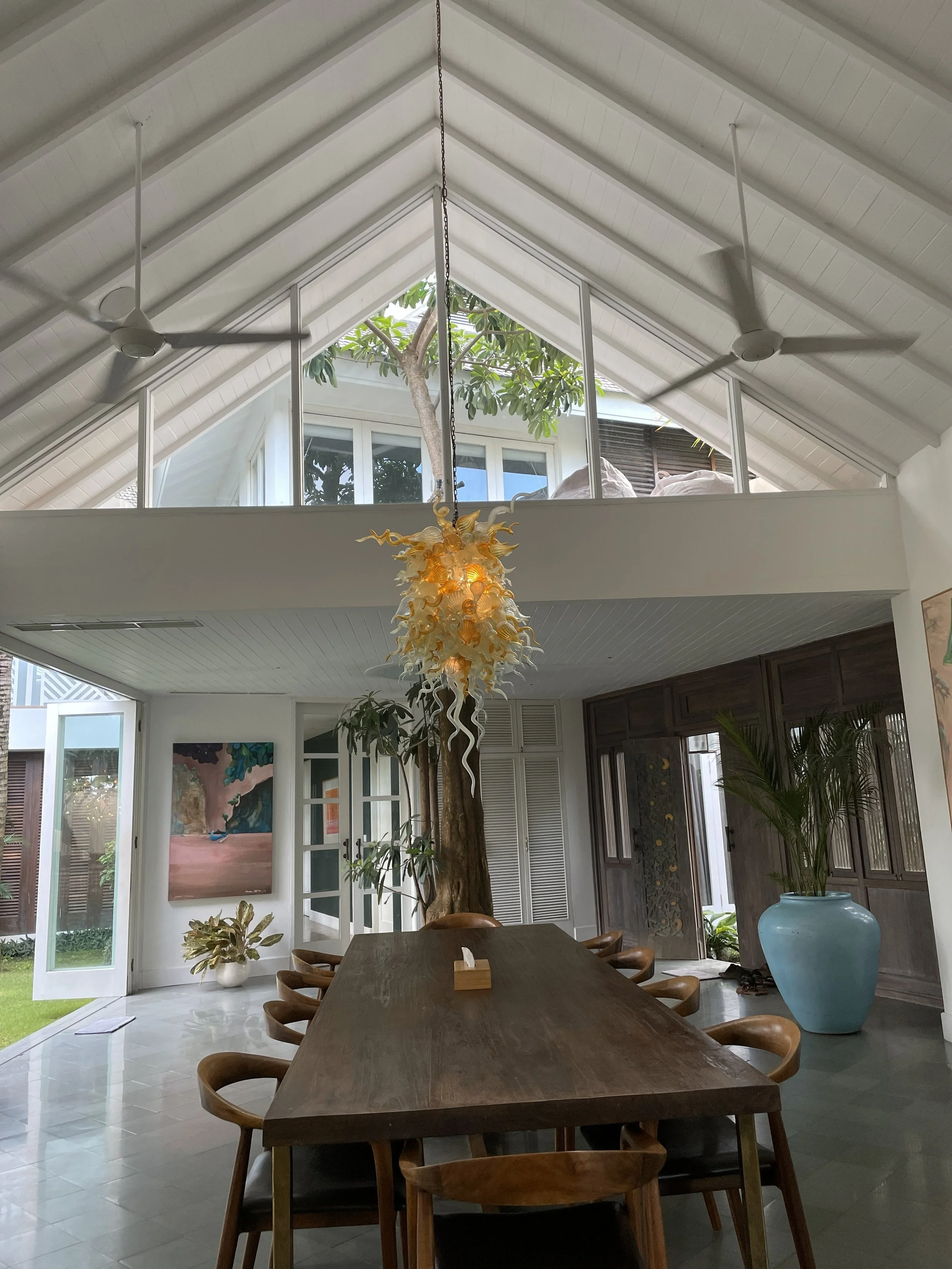 Modern dining room with a long wooden table surrounded by wooden chairs, large potted plants, an abstract painting on the wall, a unique hanging chandelier, ceiling fans, and large windows with a view of trees outside.