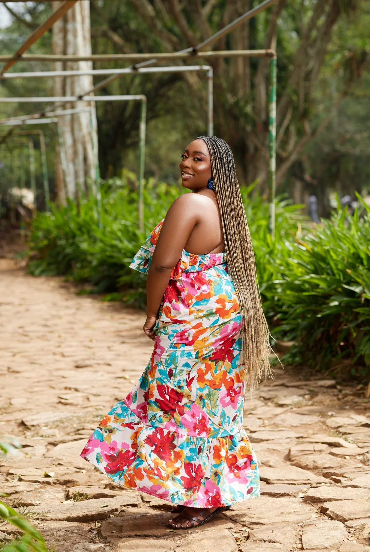 A woman in a colorful floral dress standing on a stone path in a lush green outdoor setting, smiling and looking over her shoulder.