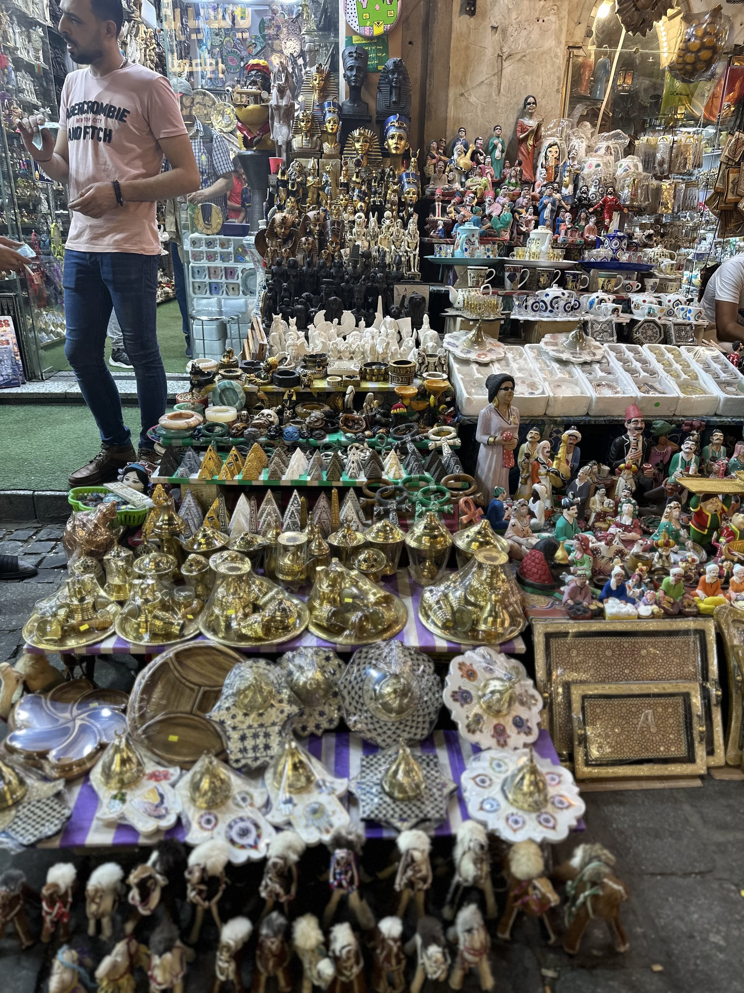 Street market stall displaying various decorative items, souvenirs, figurines, and artifacts across multiple shelves and tables.