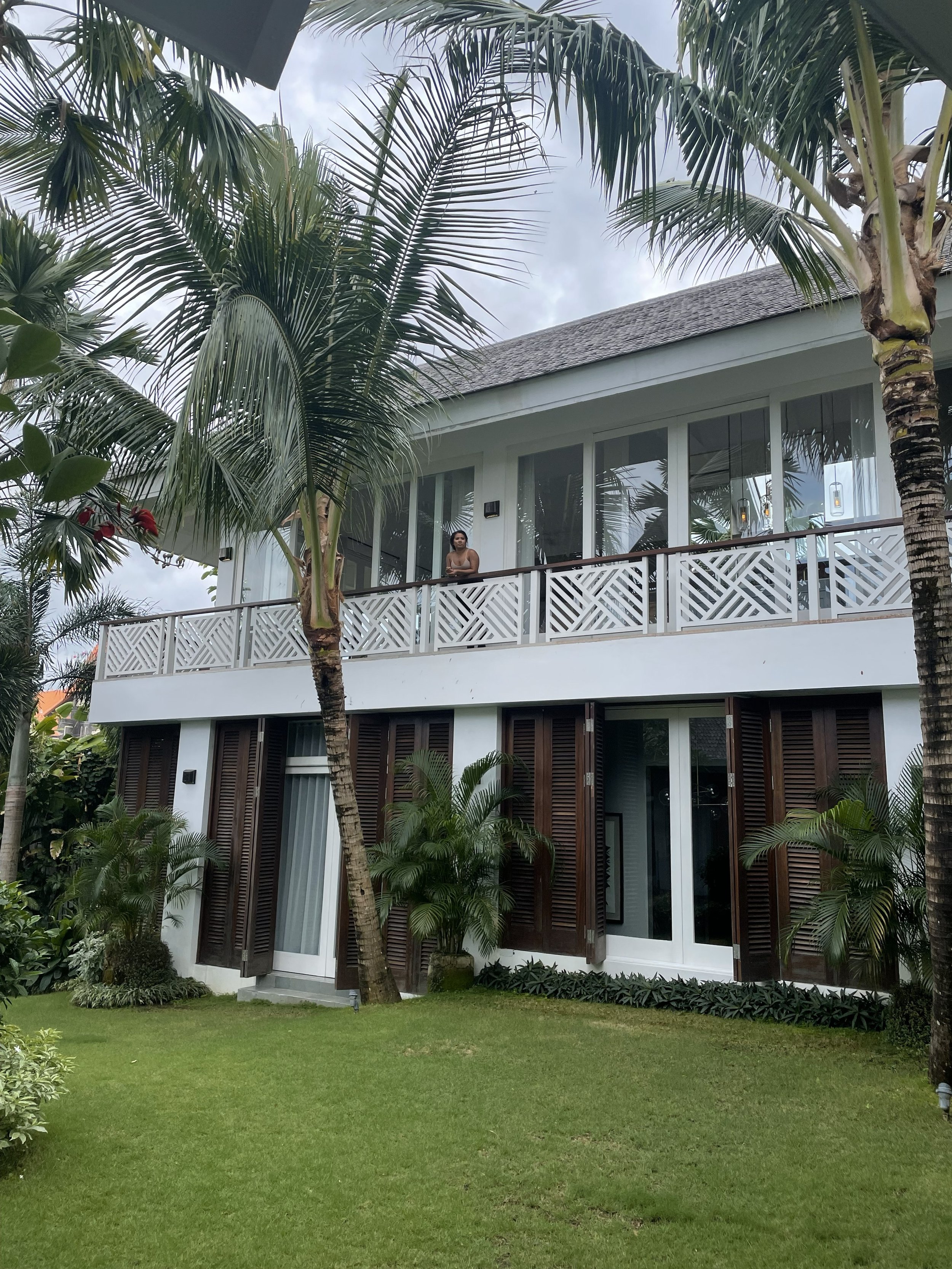 A two-story modern house surrounded by palm trees and lush green lawn, with a person standing on the upper balcony looking outward.