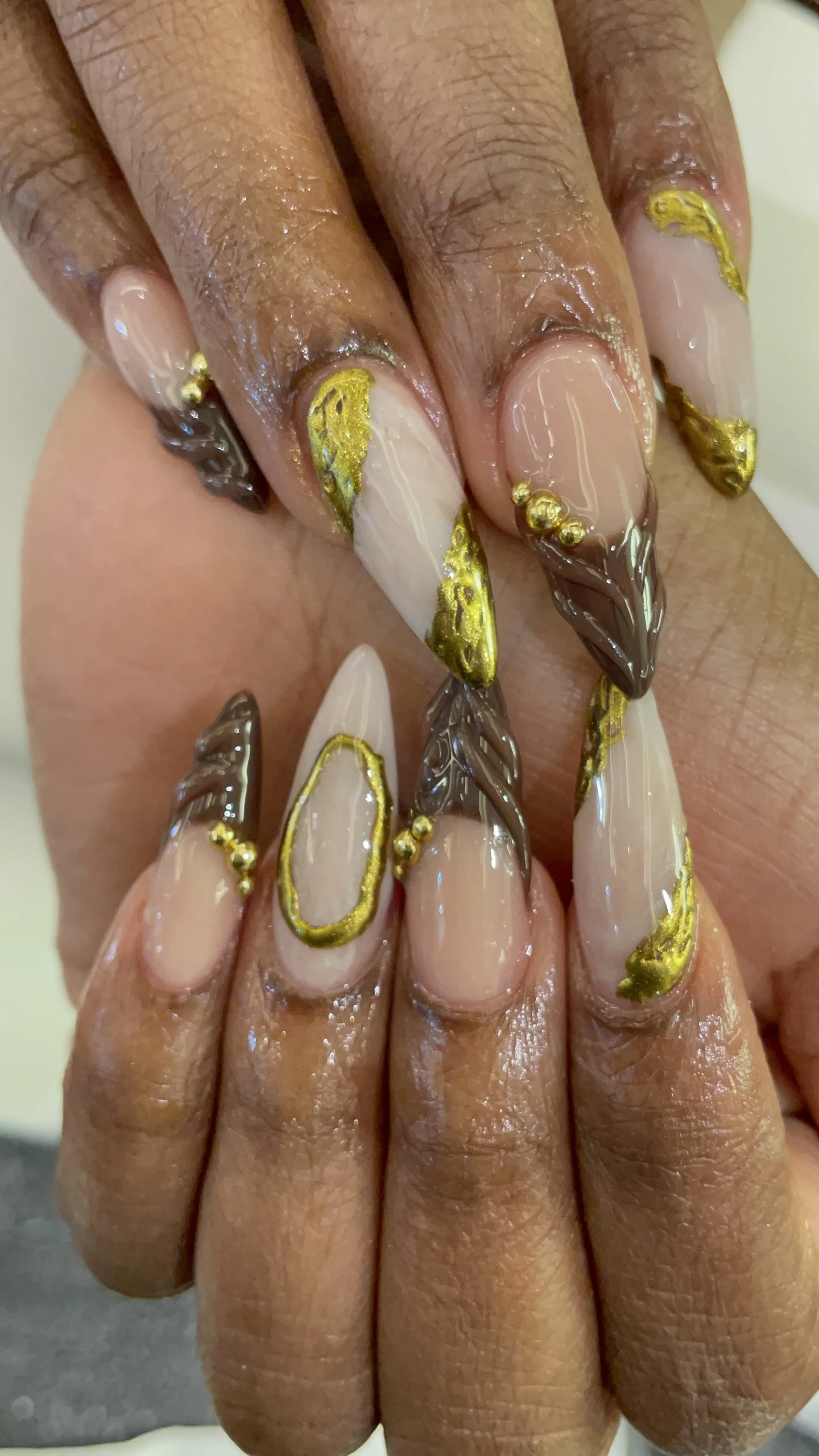 Close-up of hands with long, decorated acrylic nails in beige, gold, and dark brown with textured designs and gold embellishments.