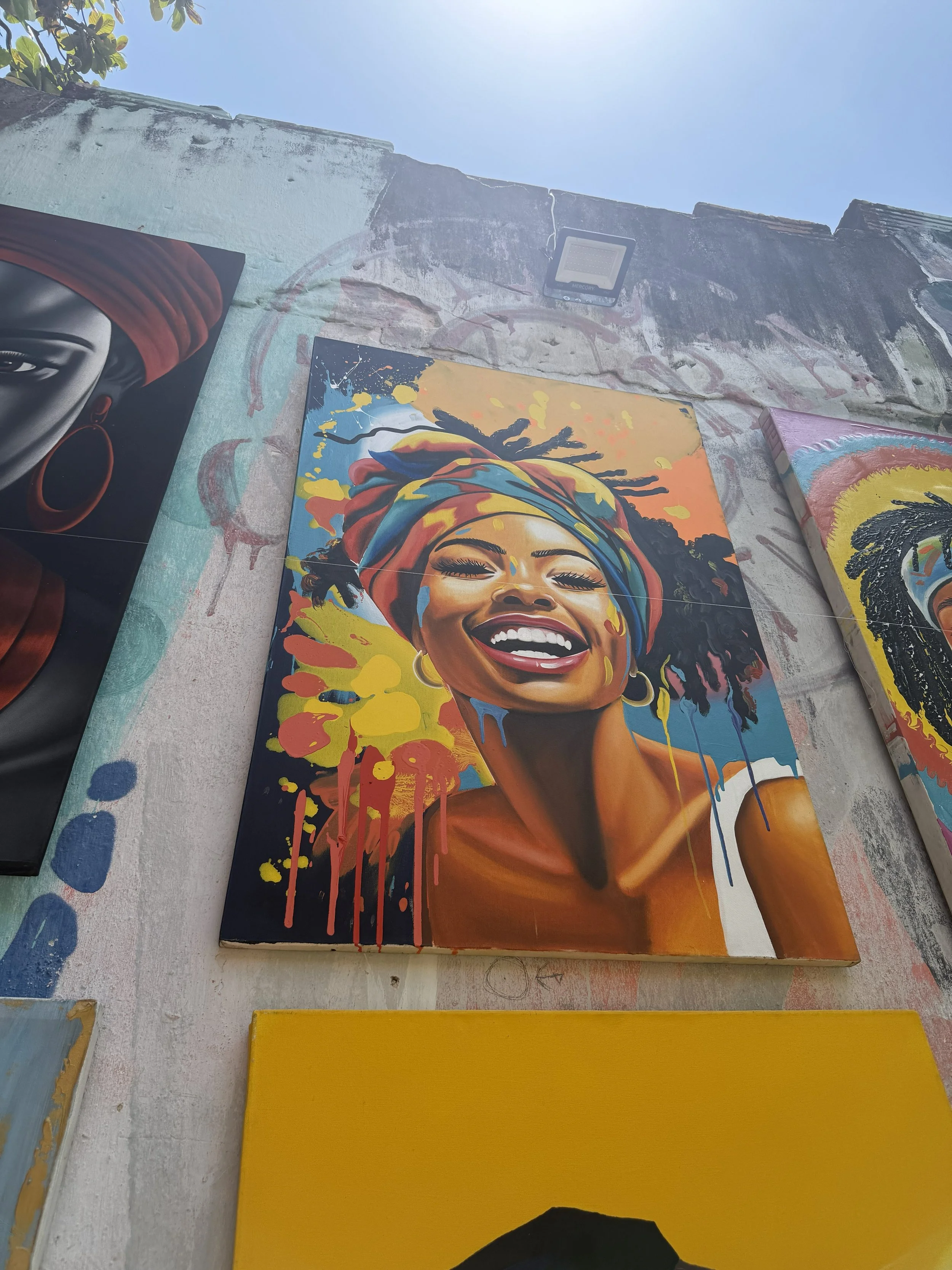 Colorful painting of a smiling woman with dreadlocks, wearing a headwrap and hoop earrings, depicted in a vibrant, abstract style with splashes of yellow, red, blue, and orange.