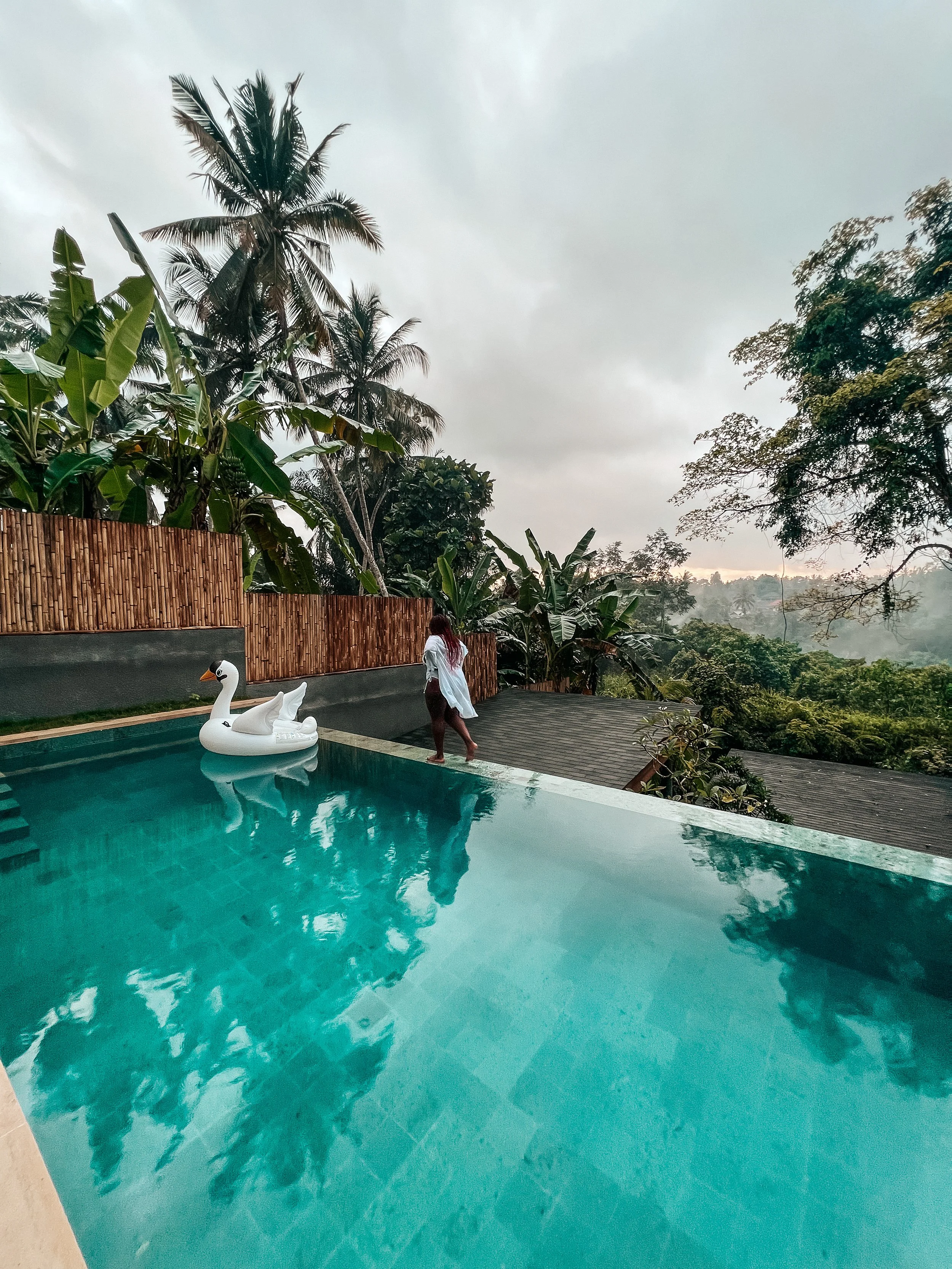 Latrice in a white swimsuit walking along the edge of a swimming pool with a swan-shaped float, surrounded by tropical trees and cloudy sky.