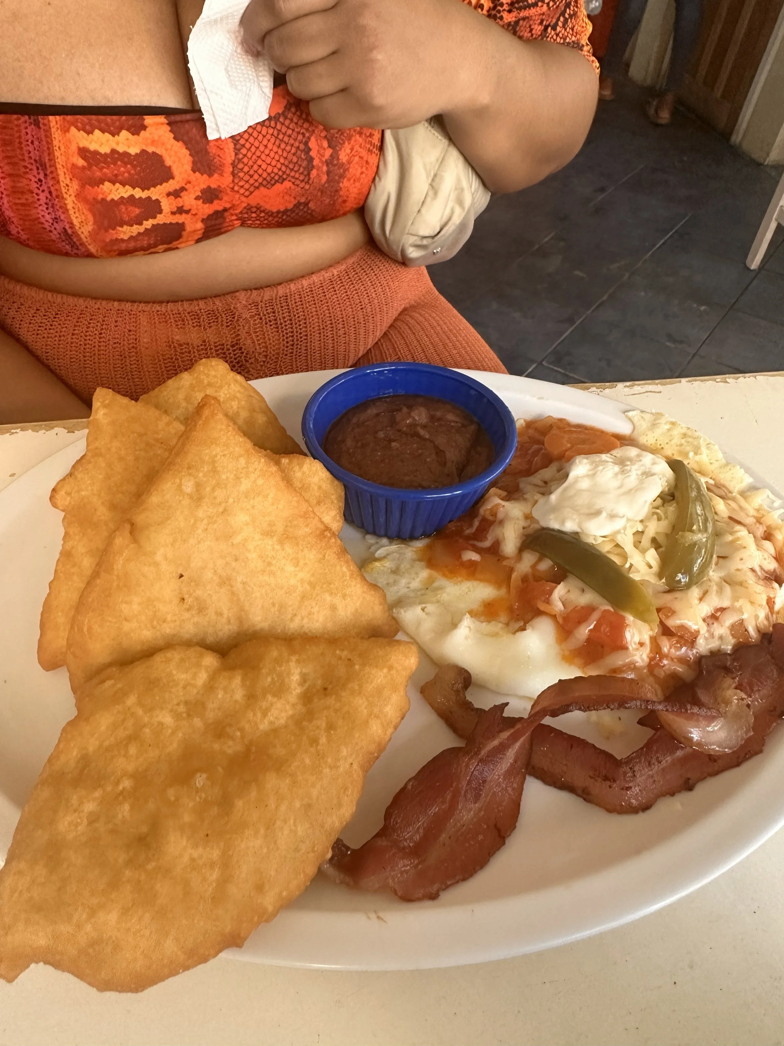 A plate of Mexican breakfast food with two fried tortilla chips, a small bowl of red salsa, a fried egg topped with tomatoes, cheese, and green peppers, and several strips of bacon. The person sitting at the table is wearing an orange shirt with a sn