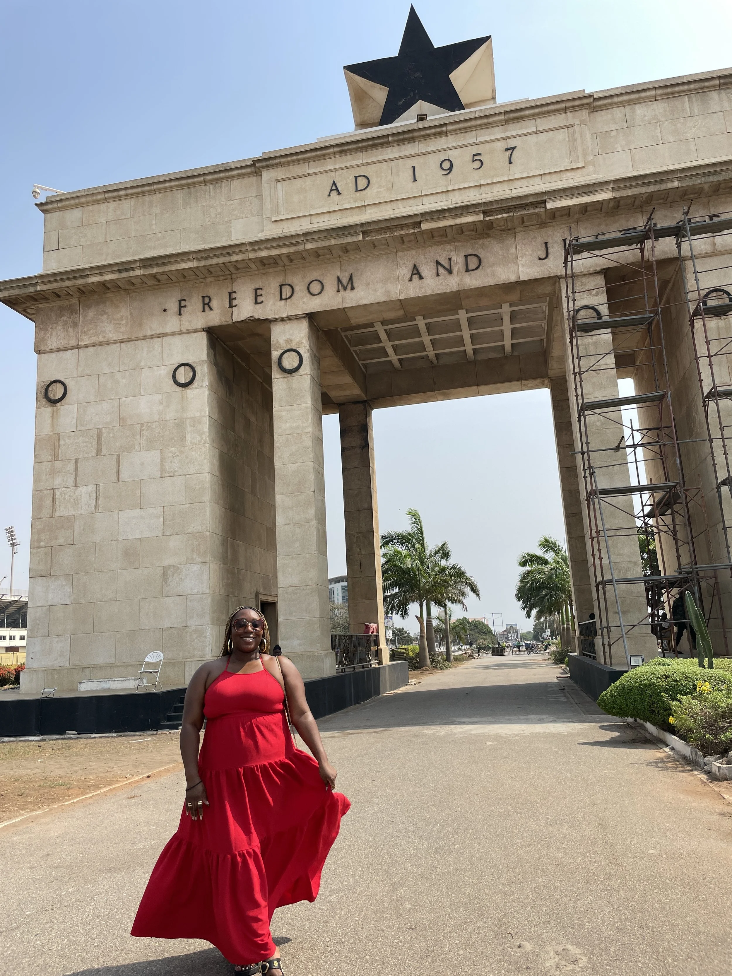 Latrice in a red dress standing in front of the Independence Arch with a large black star on top, palm trees, and a clear sky in the background.