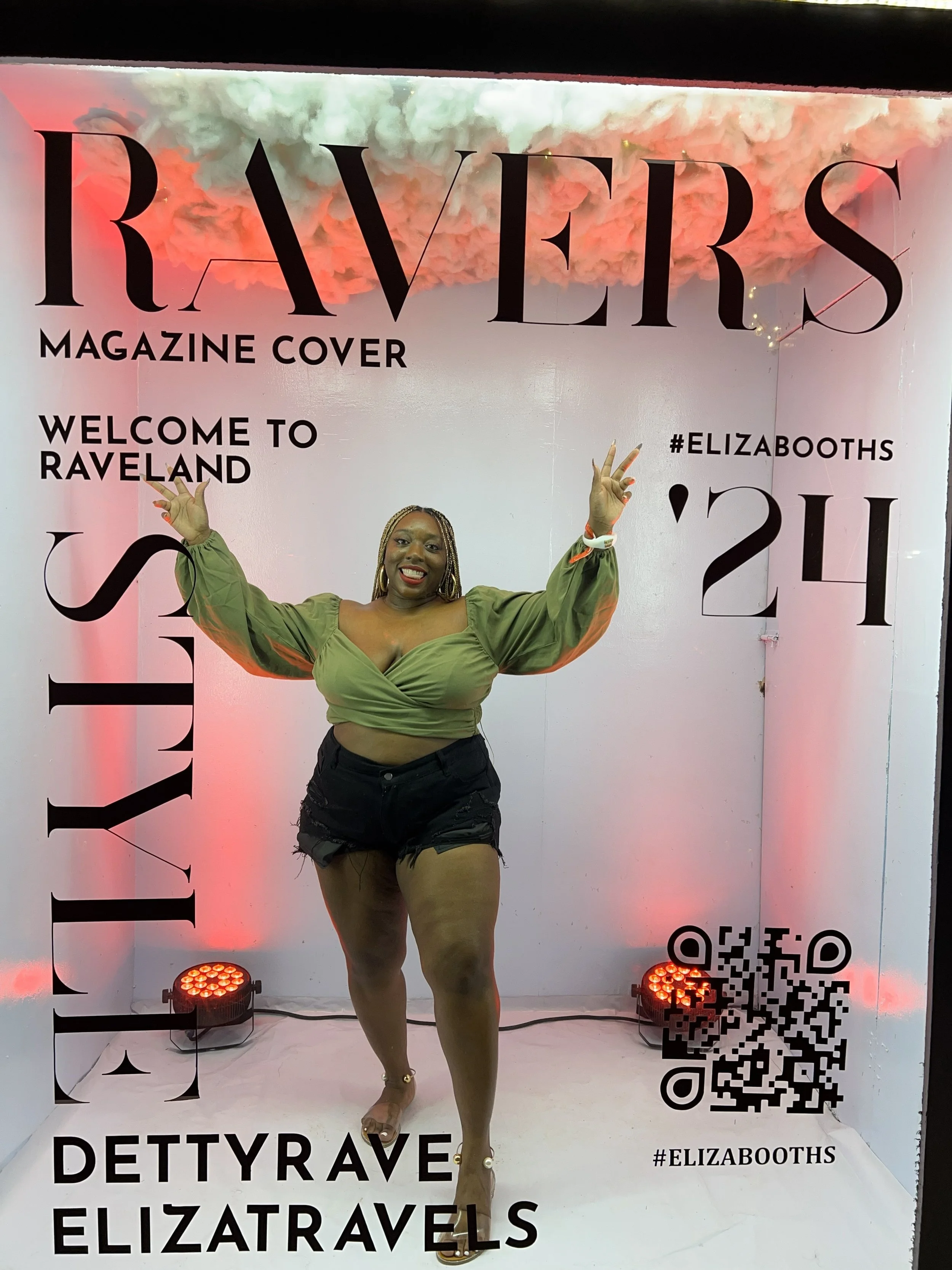Latrice standing inside a pink and white Instagram photo booth with the text 'RAVERS MAGAZINE COVER WELCOME TO RAVELAND 24' and a QR code, smiling and posing with crossed arms, with pink cloud and lighting effects in the background.