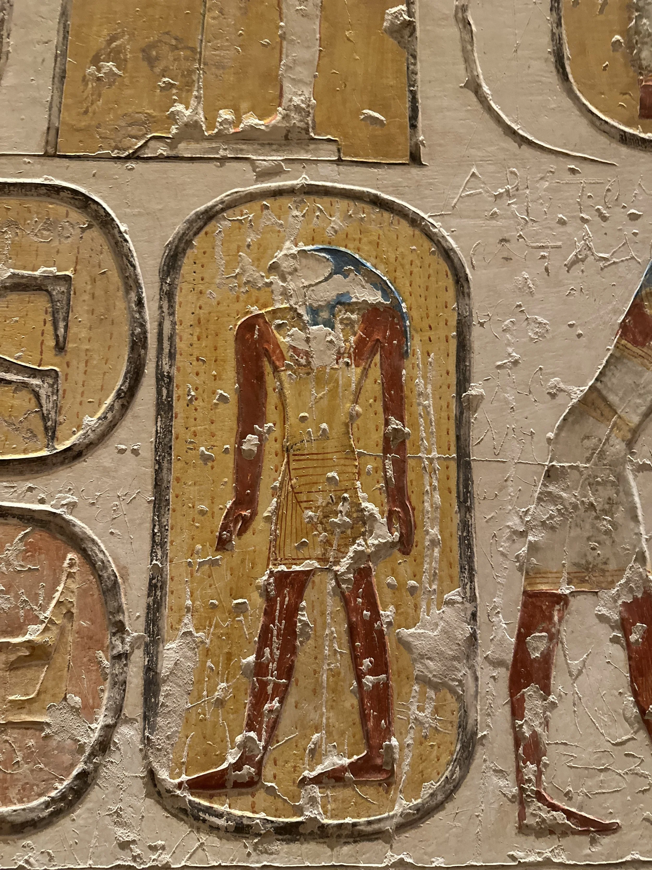 Close-up of an ancient Egyptian painted artifact depicting a human figure with a blue head, orange arms, and red legs, on a yellow background with weathered surface and chipping paint.