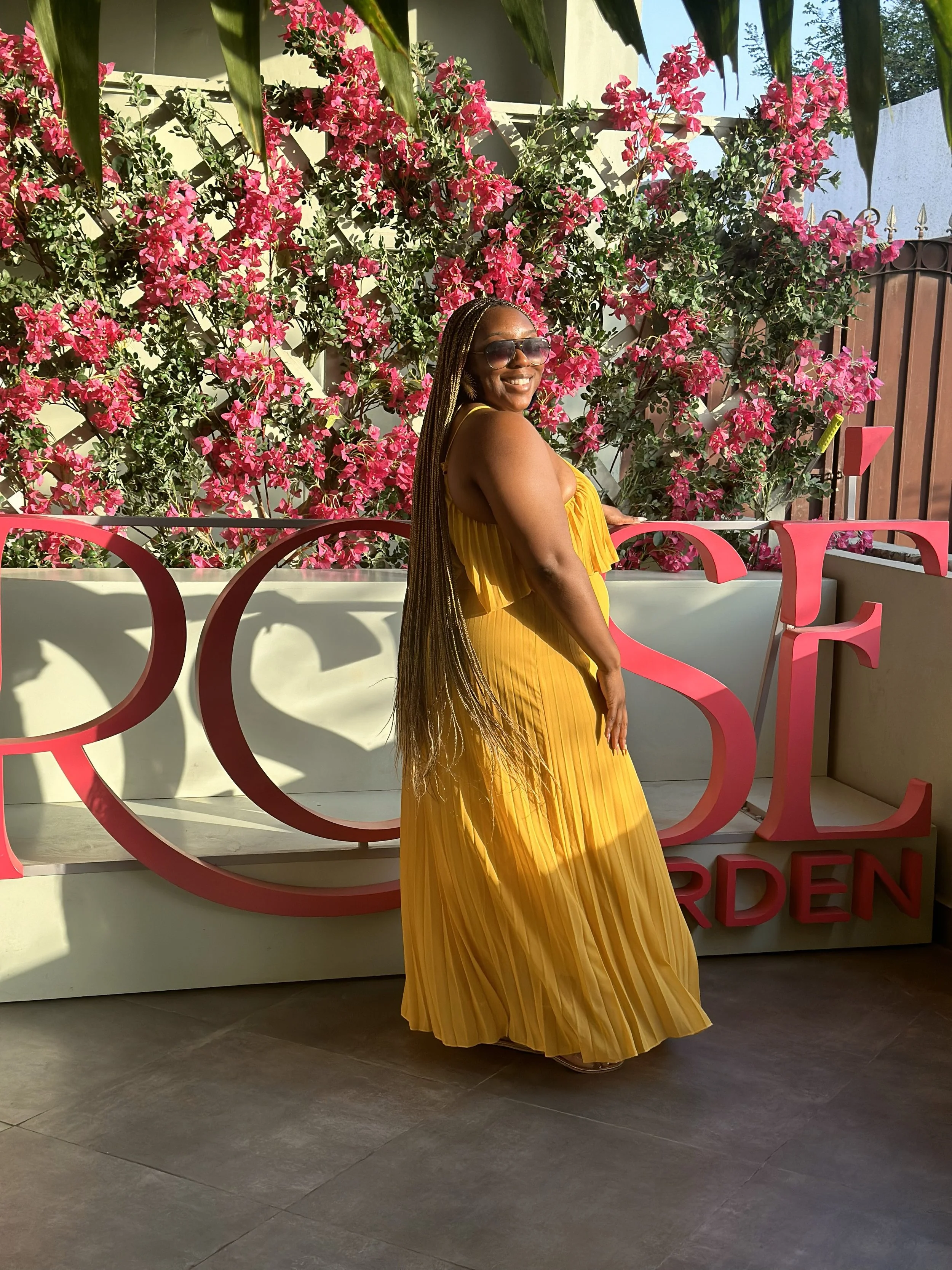 Latrice in a yellow pleated maxi dress and sunglasses smiling while standing in front of a floral backdrop with pink flowers and a sign that reads 'ROSE'.