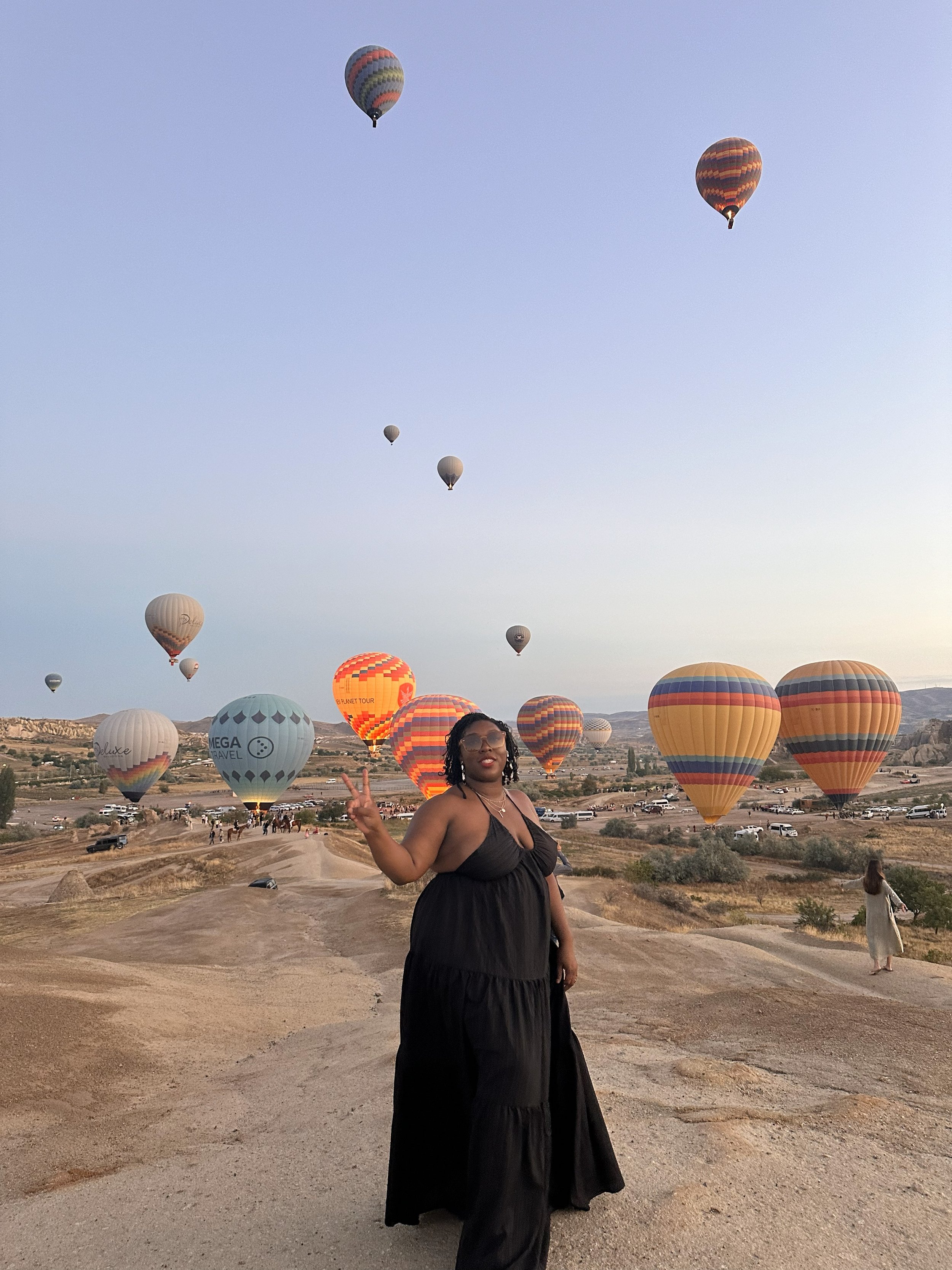 Latrice in a black dress making a peace sign in front of numerous hot air balloons in the sky at sunrise in a desert landscape.