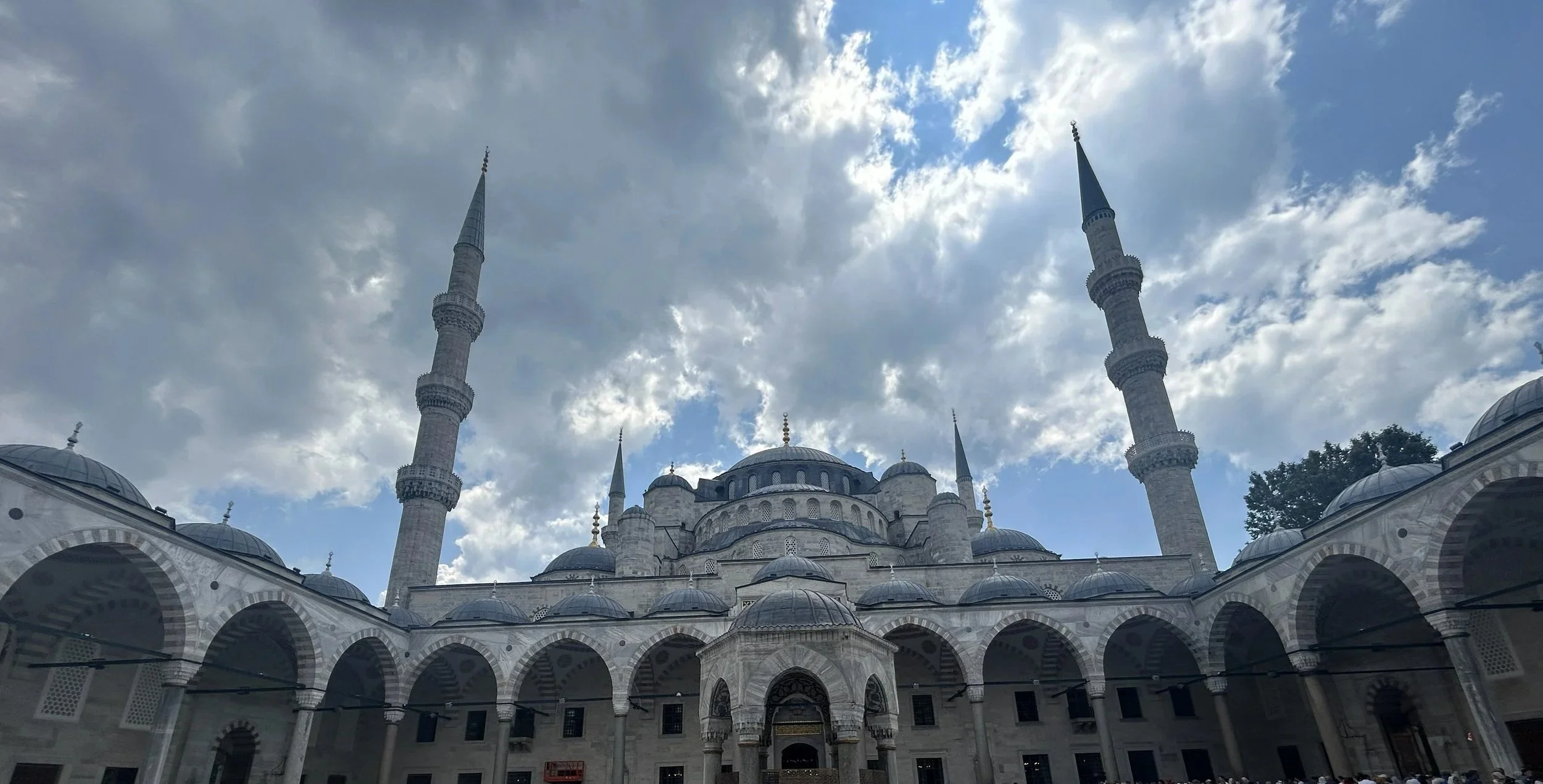 The image shows a grand mosque with multiple domes and four tall minarets against a partly cloudy sky.