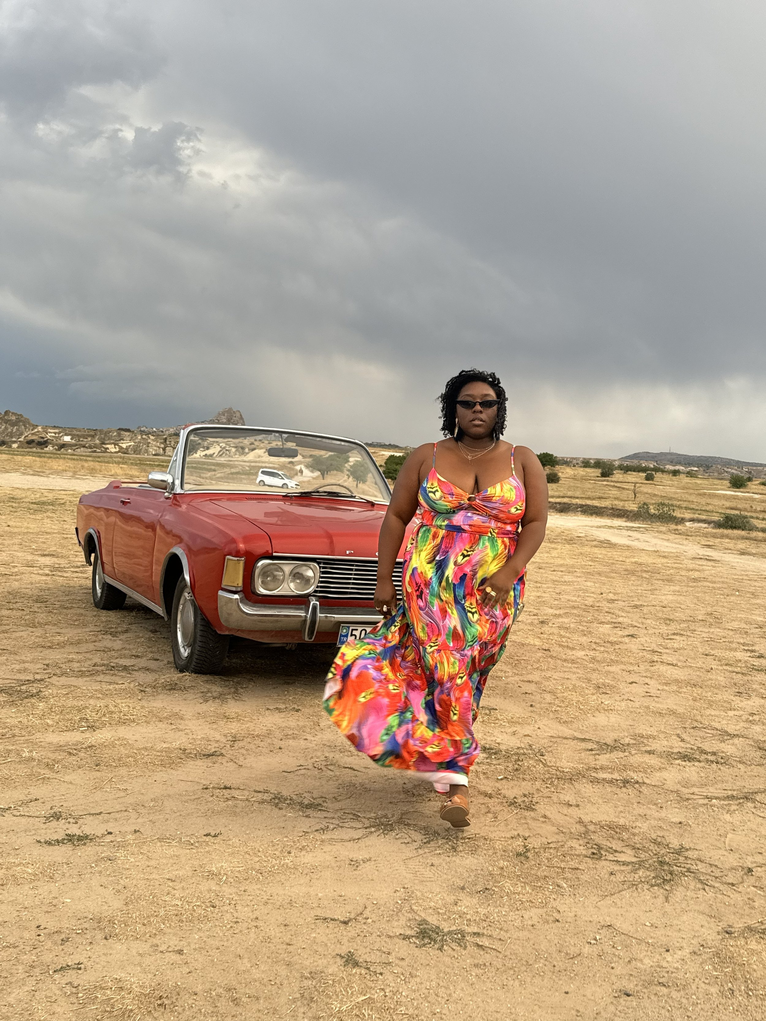 Latrice in a colorful flowing dress walking in a desert with a red vintage convertible car and stormy sky in the background.