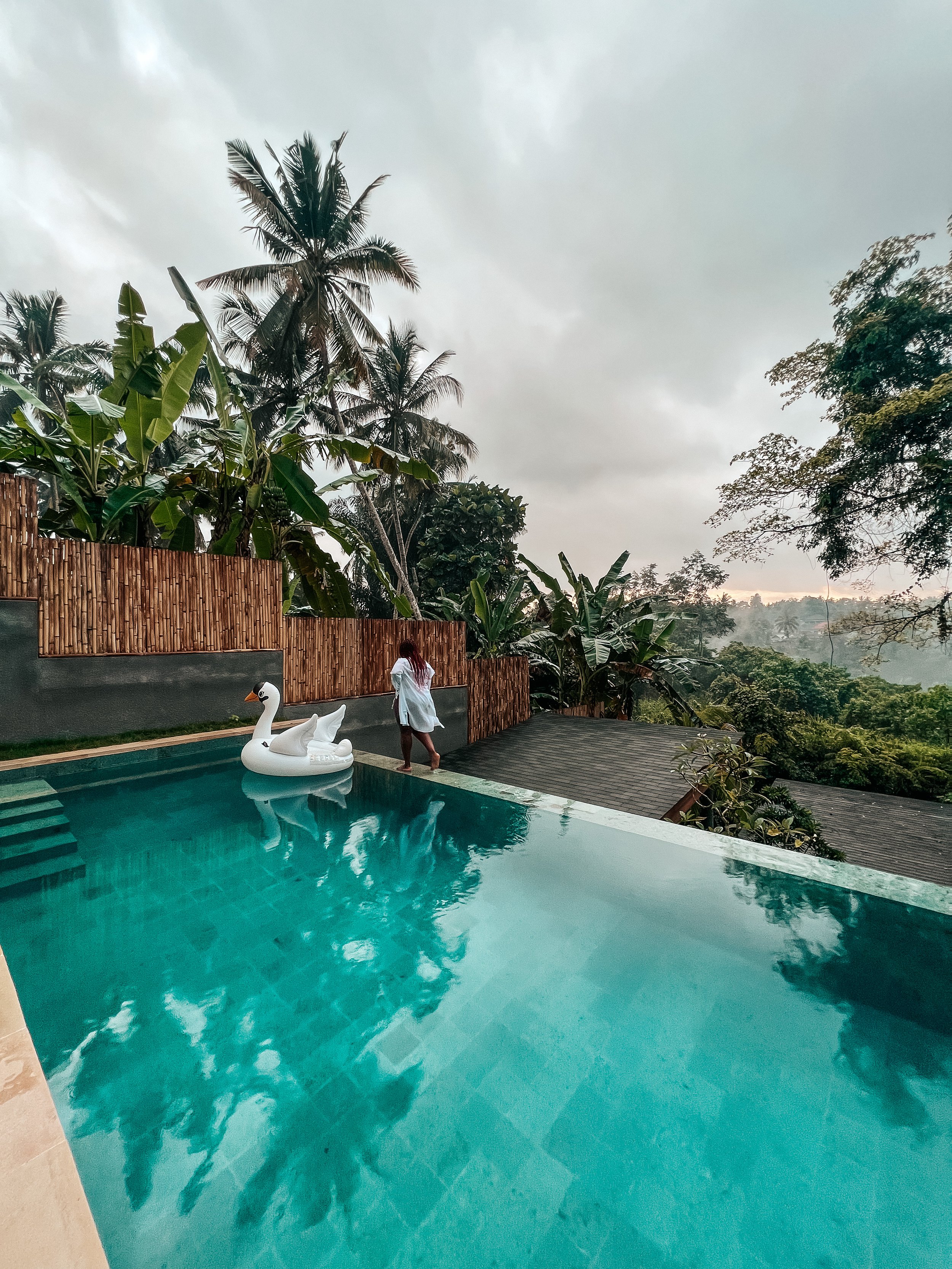Latrice in a white dress standing by a swimming pool with a swan-shaped float, surrounded by tropical trees under a cloudy sky.