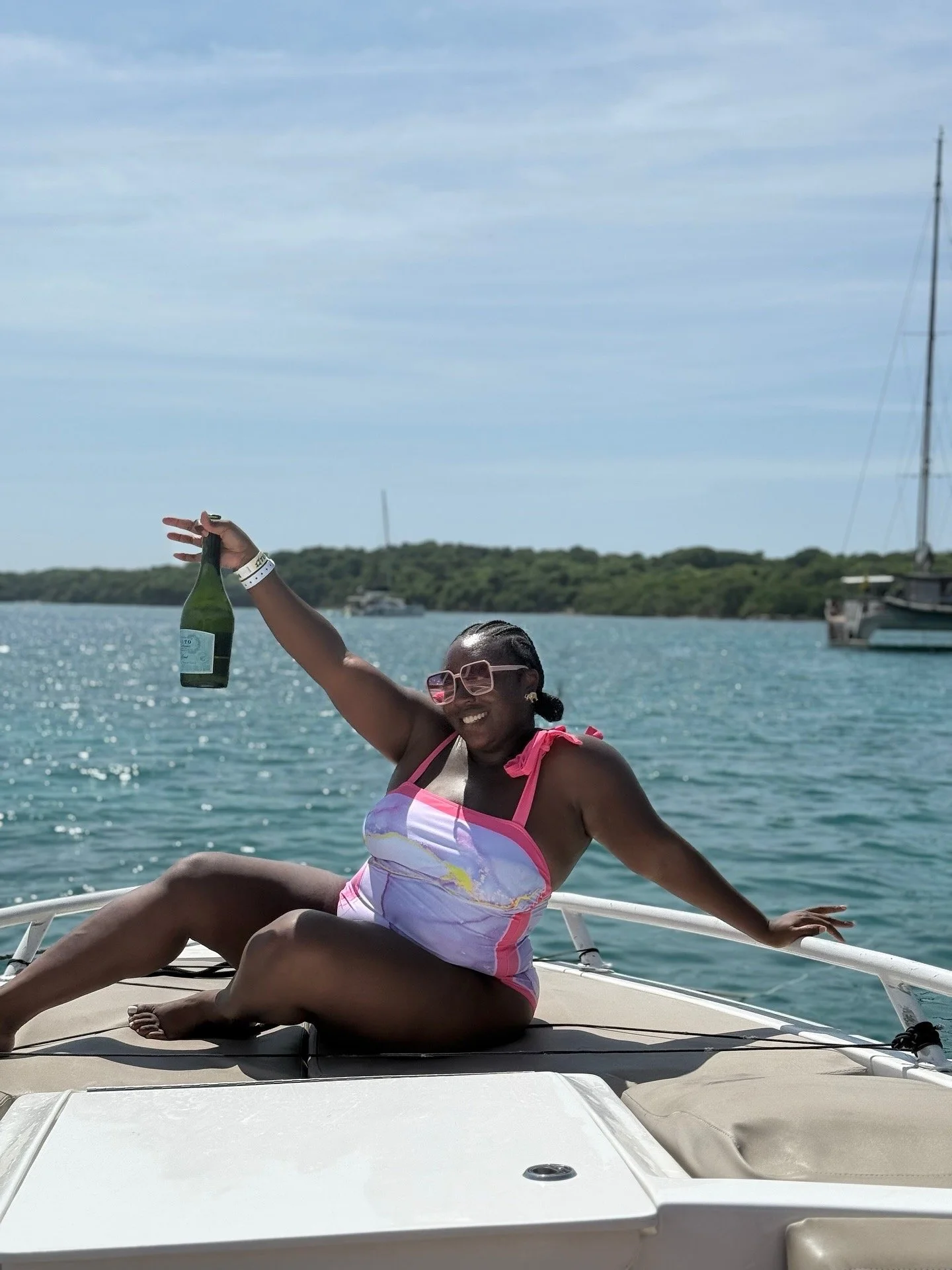 Latrice in a colorful swimsuit sitting on a boat, holding a bottle of champagne, smiling, with water and sailboats in the background.