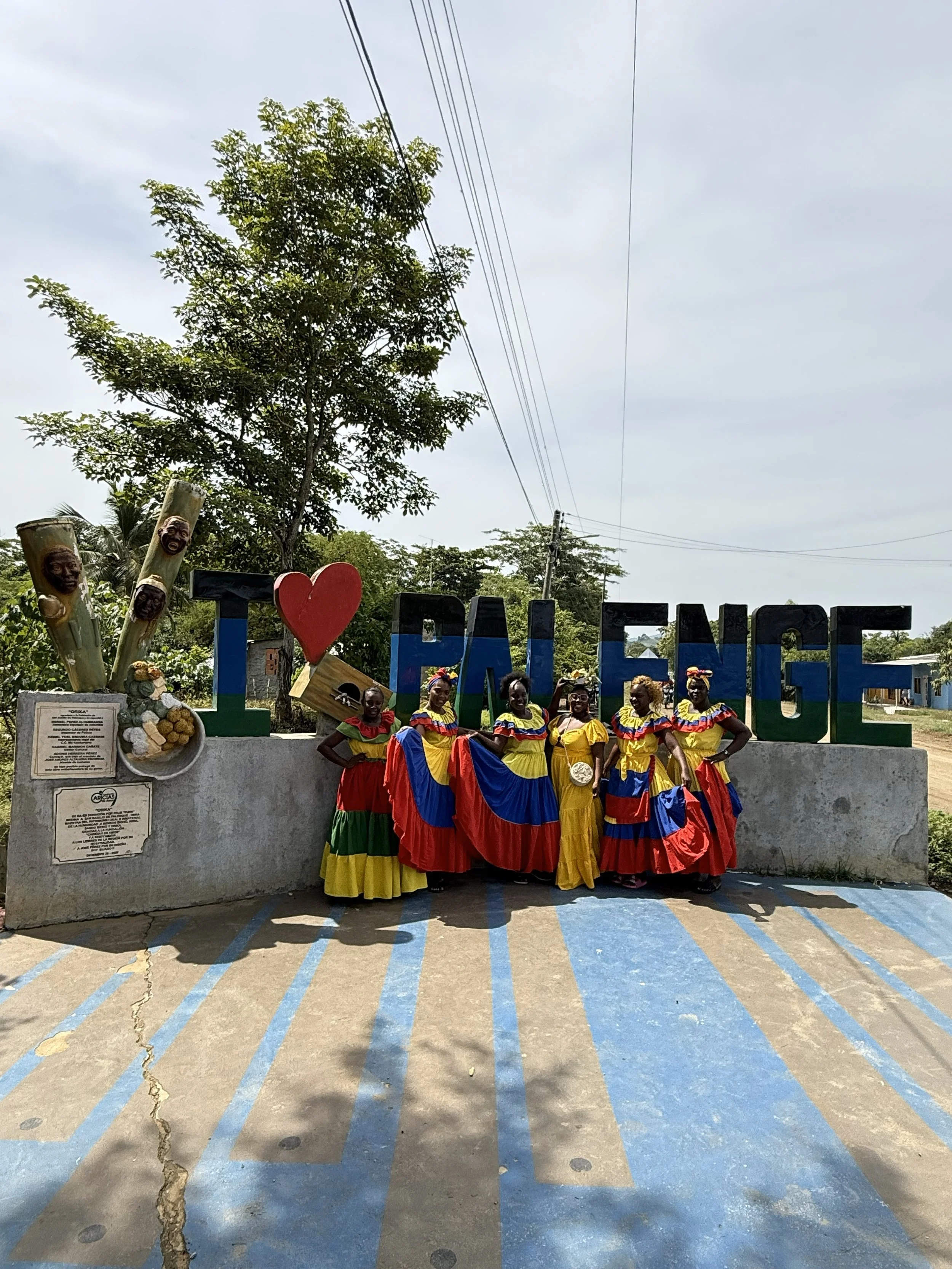 Group of women in colorful traditional dresses standing in front of a sign that reads 'I ❤️ PALENGE', with trees and power lines in the background.