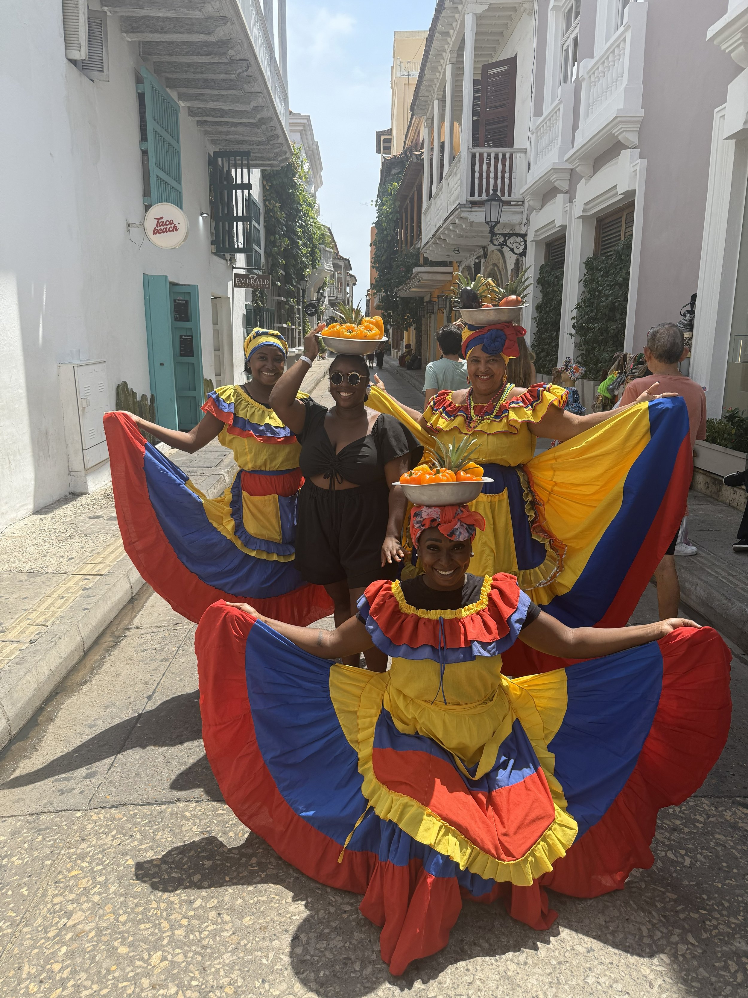 Four women dressed in colorful traditional Colombian dresses with yellow, blue, and red skirts pose on a narrow street, with three women balancing bowls of fruit on their heads and one woman standing in the middle smiling.