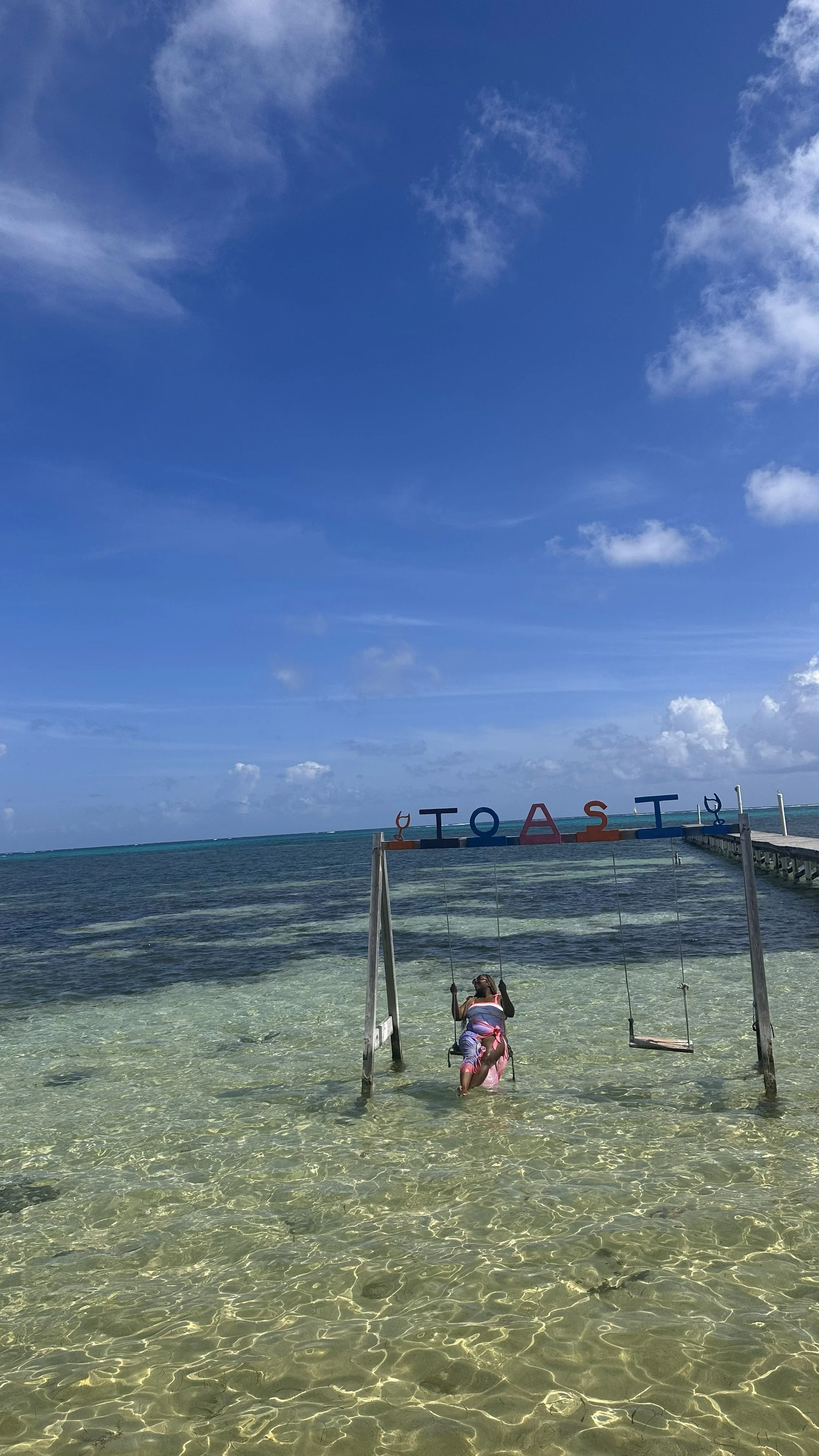 Latrice playing on a swing set in shallow clear ocean water near a pier, with a bright blue sky and scattered clouds in the background.