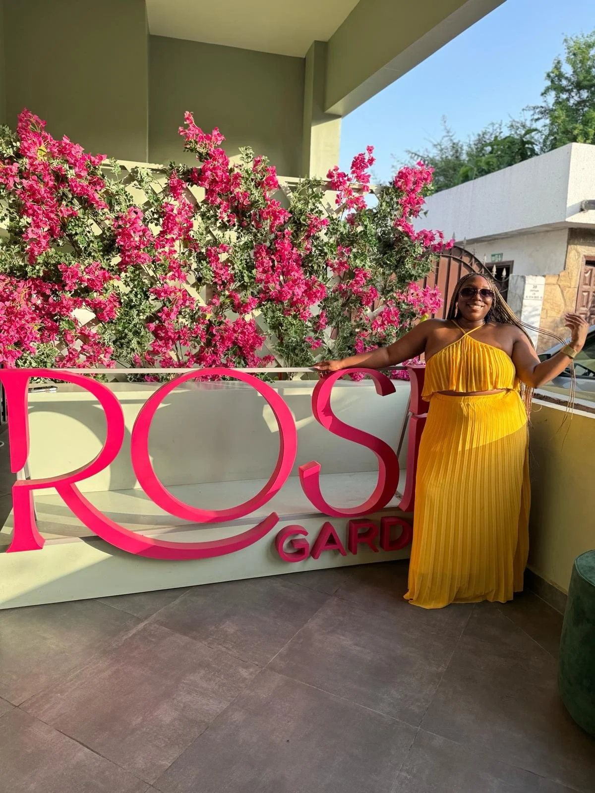 Latrice standing next to a pink and white sign that reads 'ROS GARD,' with vibrant pink bougainvillea flowers in the background, on a balcony. She is wearing a yellow pleated dress, sunglasses, and smiling.