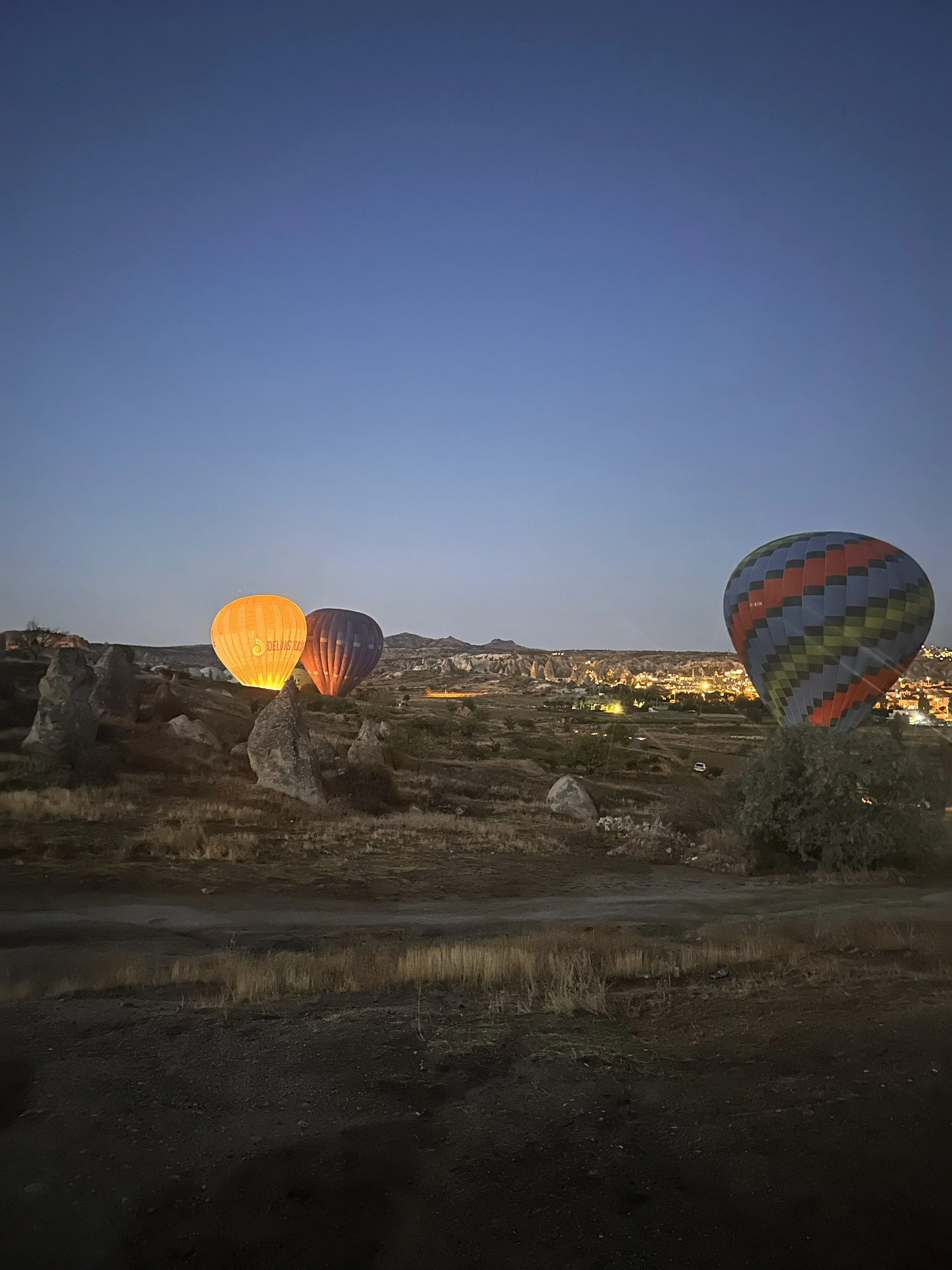 Three colorful hot air balloons floating in the early evening sky over a rocky landscape with distant city lights.