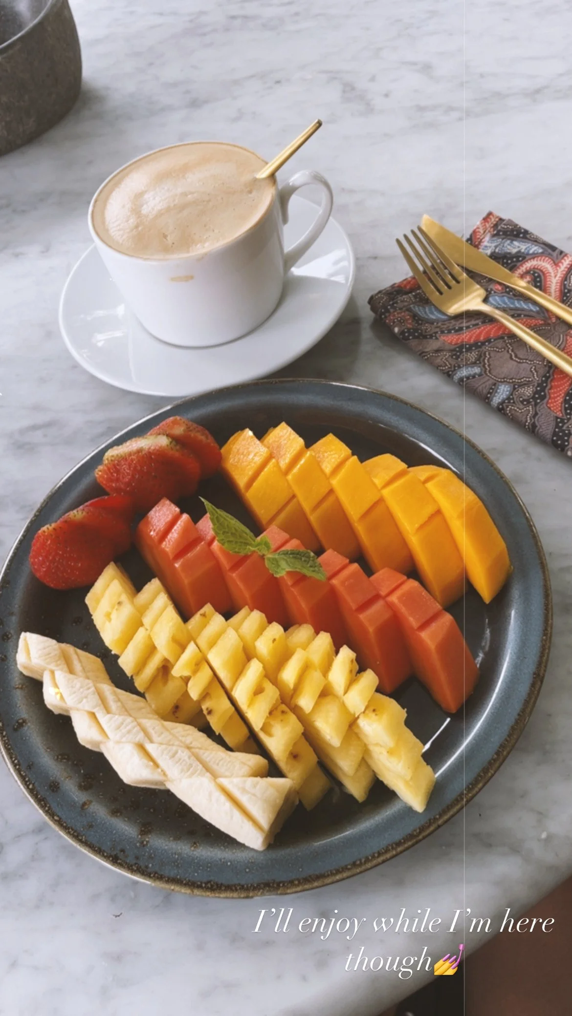 A plate of sliced tropical fruits, including strawberries, watermelon, pineapple, and mango, garnished with a mint leaf, on a marble table with a cup of coffee or tea nearby.