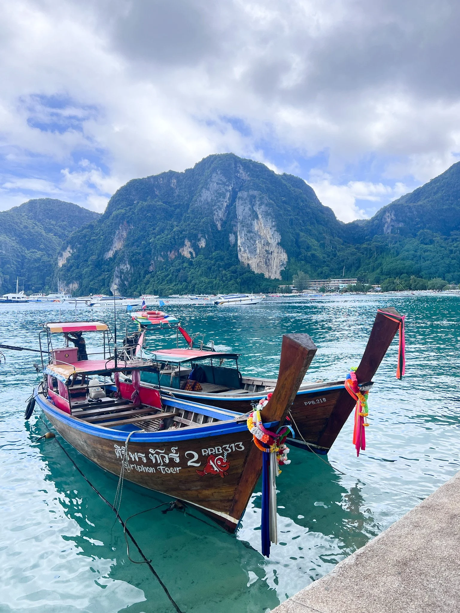 Traditional wooden boat decorated with colorful ribbons, floating near a stone pier with scenic mountain and cloudy sky in the background.