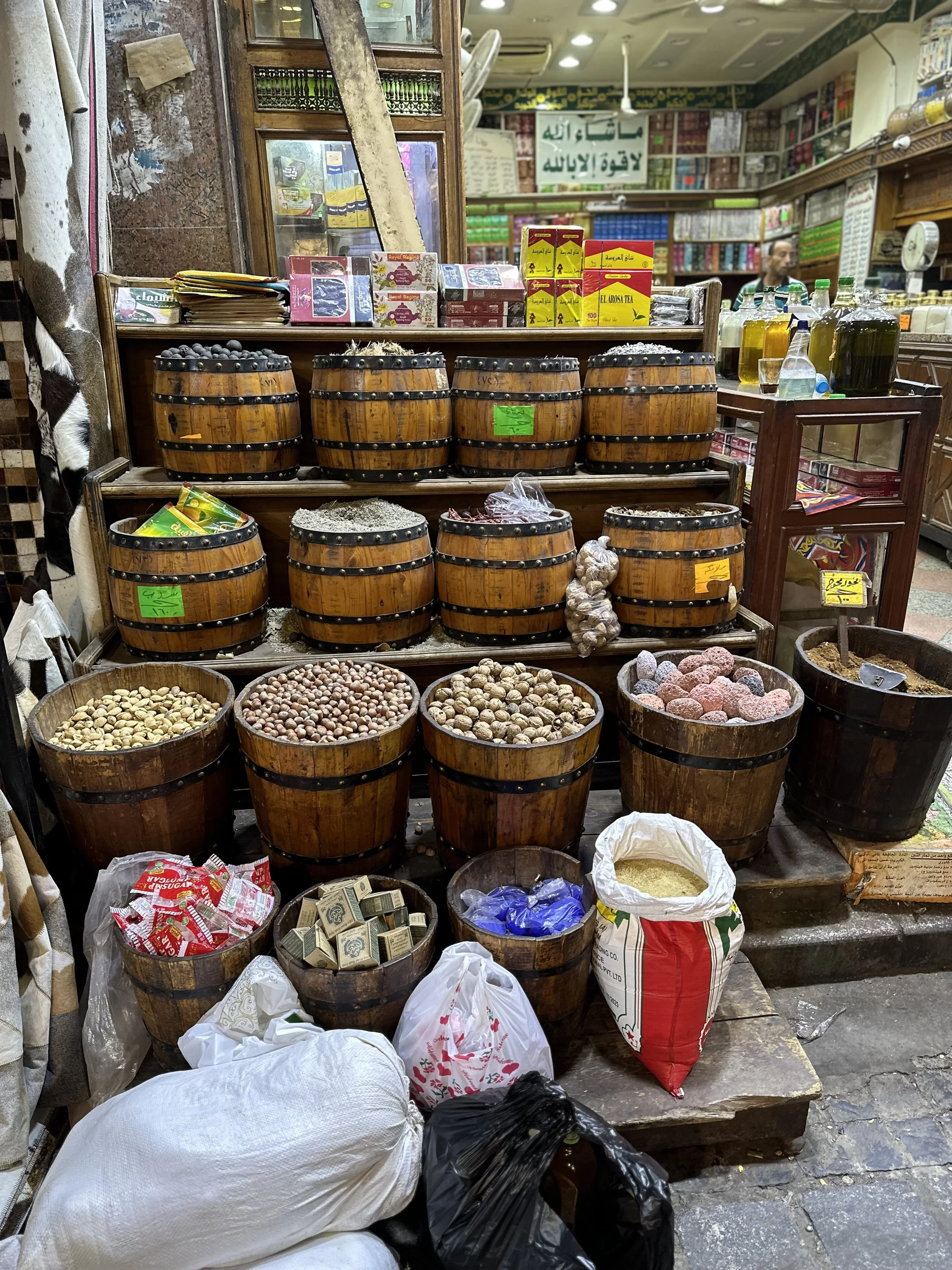 A market stall with multiple wooden barrels filled with various nuts and spices, some in bags. There are boxes of tea on the upper shelf, and bottles of liquids on a table to the right. Shelves with colorful products are visible in the background.