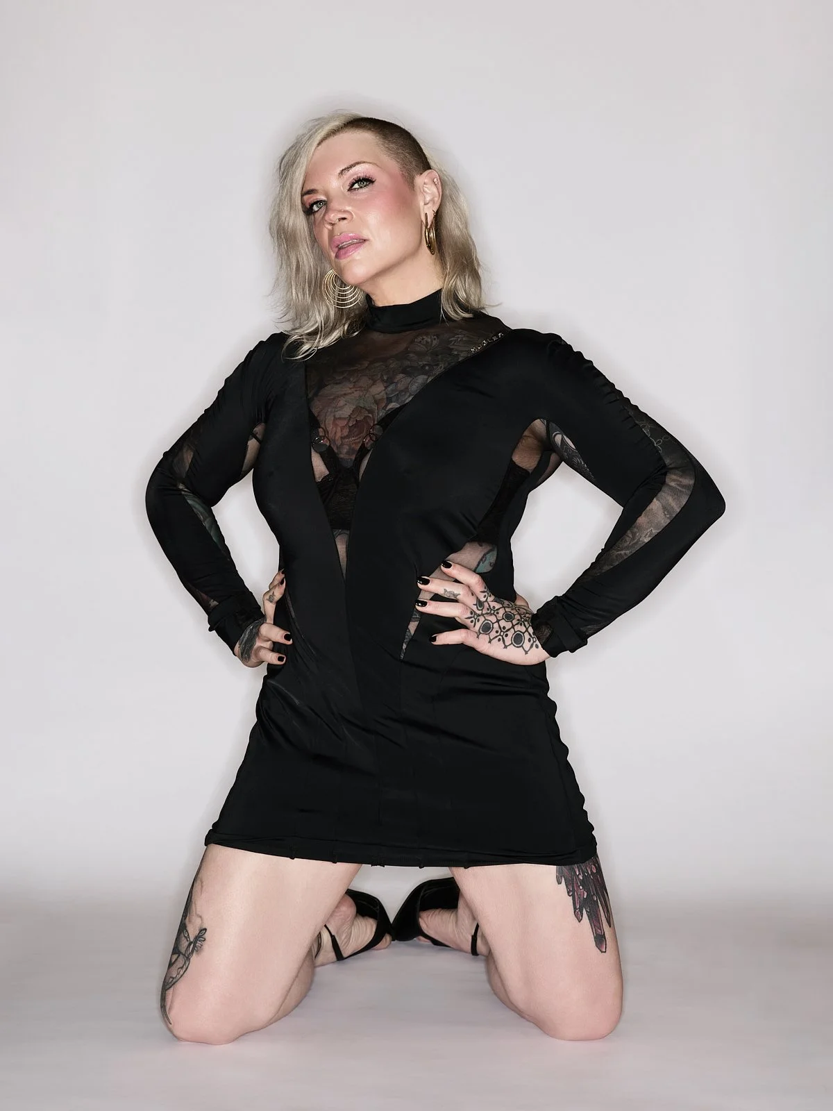 Chicago Mistress Erin Black is wearing a black long sleeve mesh dress kneeling on the ground with her hands on her hips.