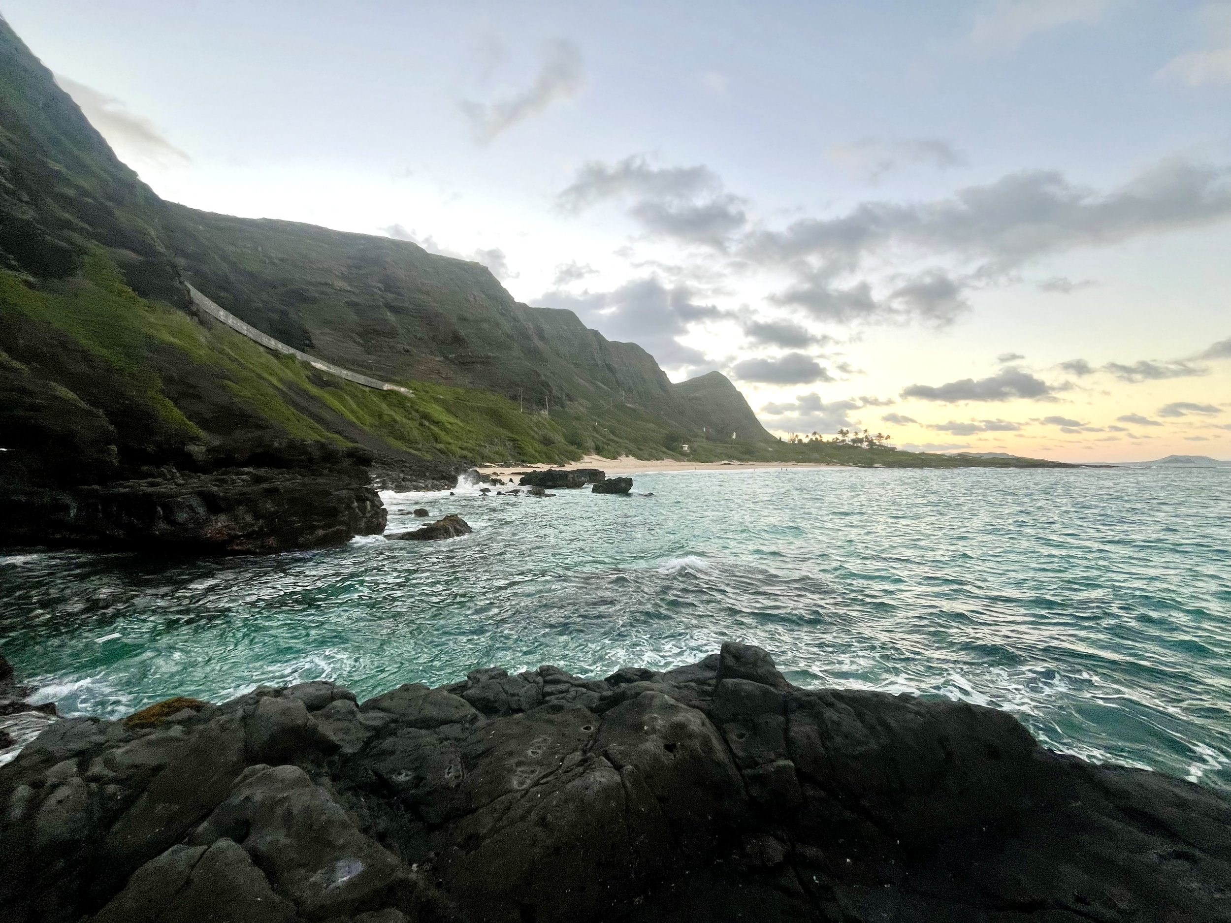Makapuu Beach coastal view showing dark rocky shore in the foreground, turquoise water, lush green mountains on the left, and a cloudy sky during sunset in the background.