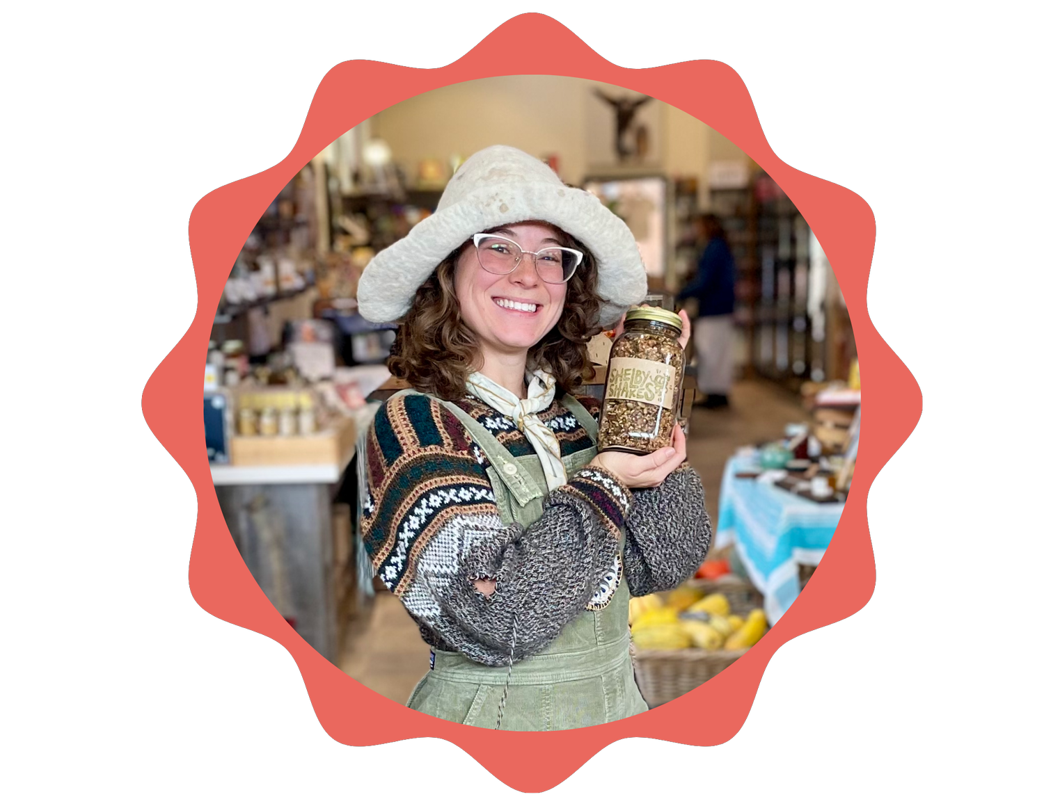 A woman smiling and holding a jar of granola in a market. She is wearing a large hat, glasses, a patterned sweater, and overalls. The background shows market stalls with produce and other shoppers.