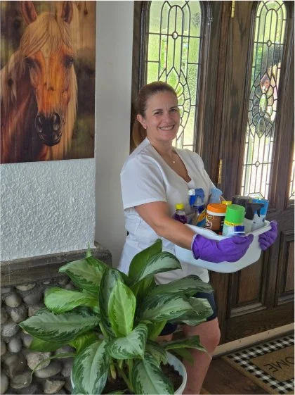 A woman smiling inside a house, holding a tray of household cleaning supplies, wearing purple rubber gloves, with decorative stained glass doors behind her and a large green plant nearby.
